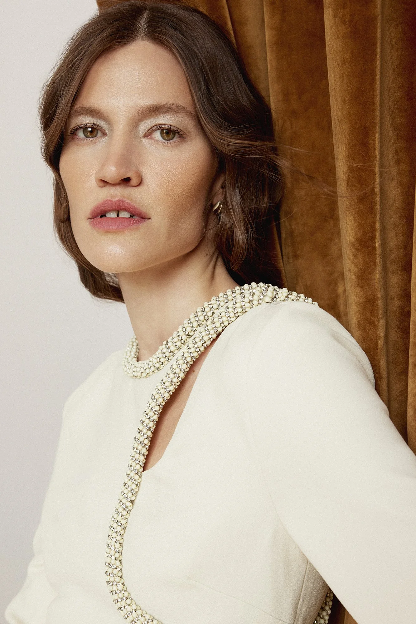 A woman with brown hair and fair skin wearing a white dress with pearl necklace and earrings, posing against a brown curtain background.