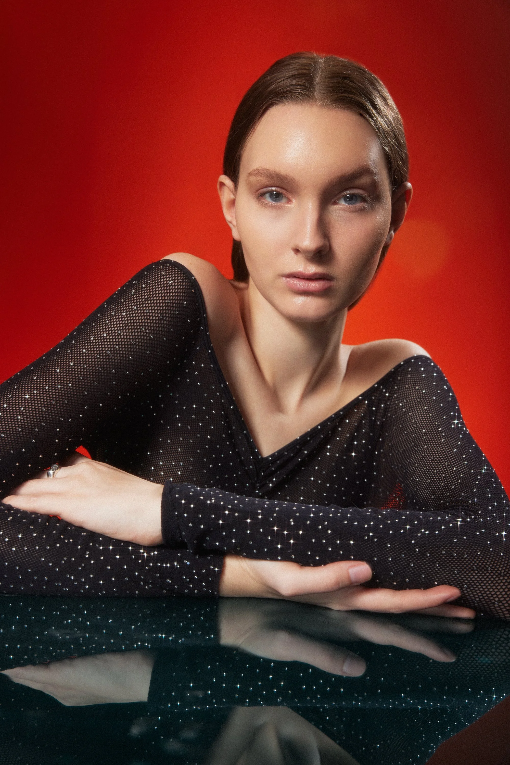 A woman with light skin and brown hair styled back, wearing a black, sheer, sparkly top, resting her arms on a reflective surface, against a red gradient background.
