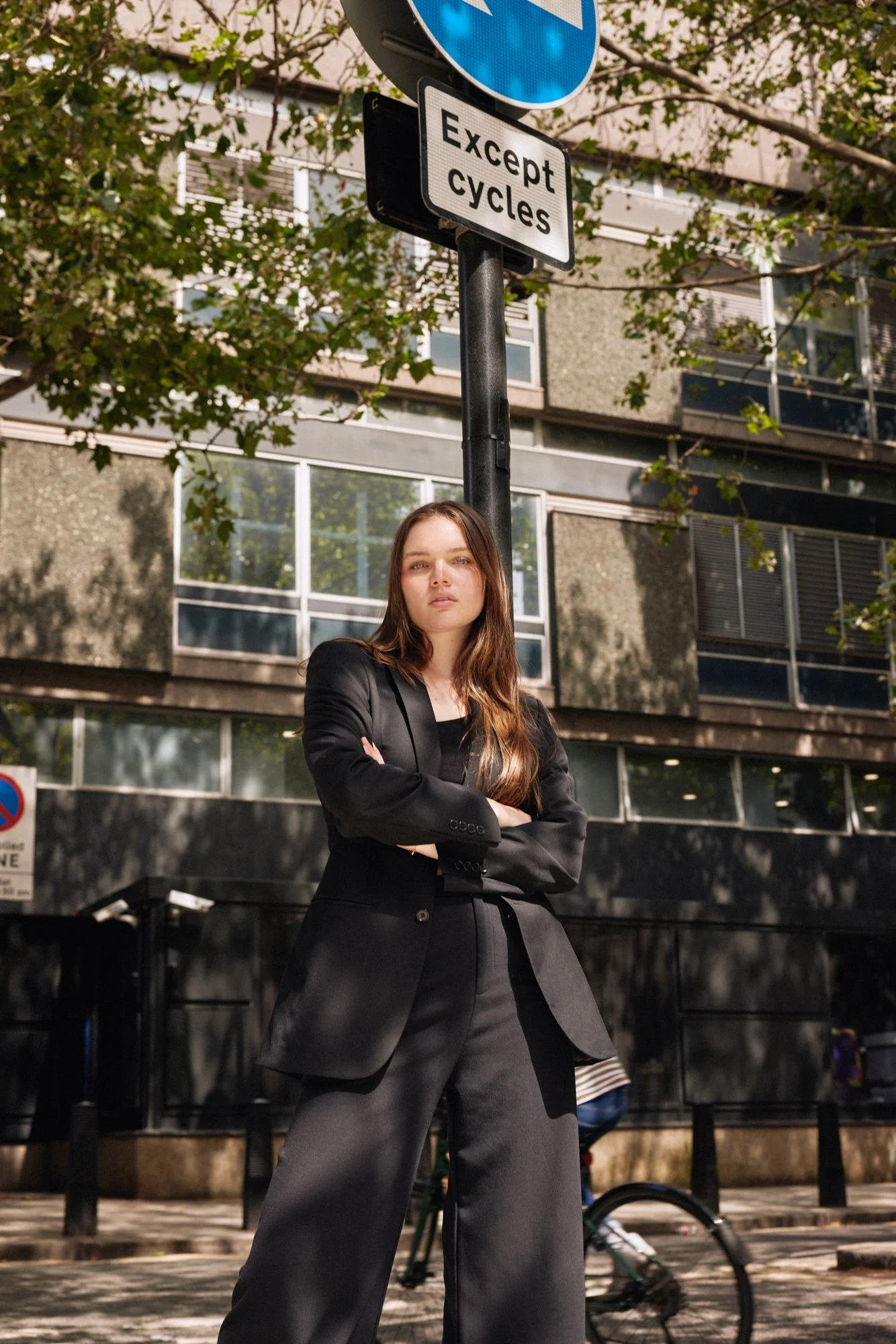 A young woman with long brown hair wearing a black blazer and black wide-leg pants stands with arms crossed in an urban street setting. There is a street sign above her that says "Except cycles." Behind her are modern buildings and trees.