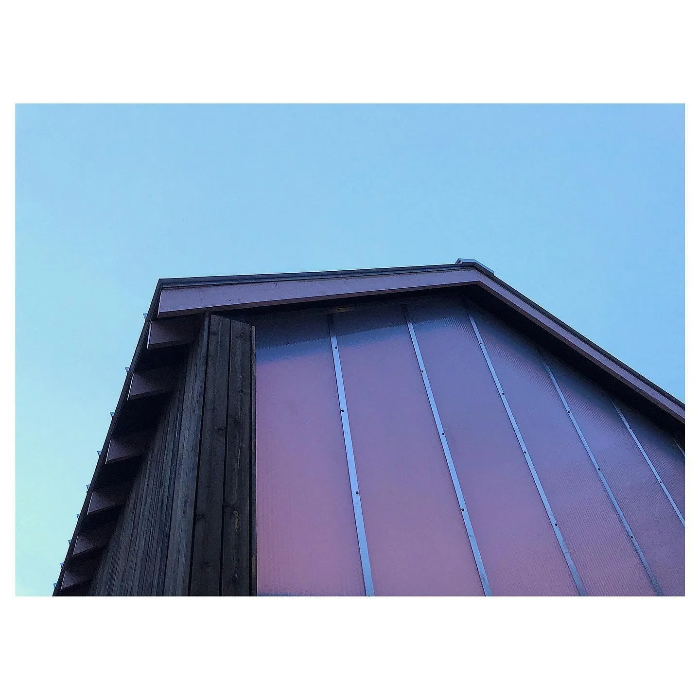 A photo taken from below of the corner of a modern building with a sloped roof, pink exterior panels, and a wooden side, against a clear blue sky.