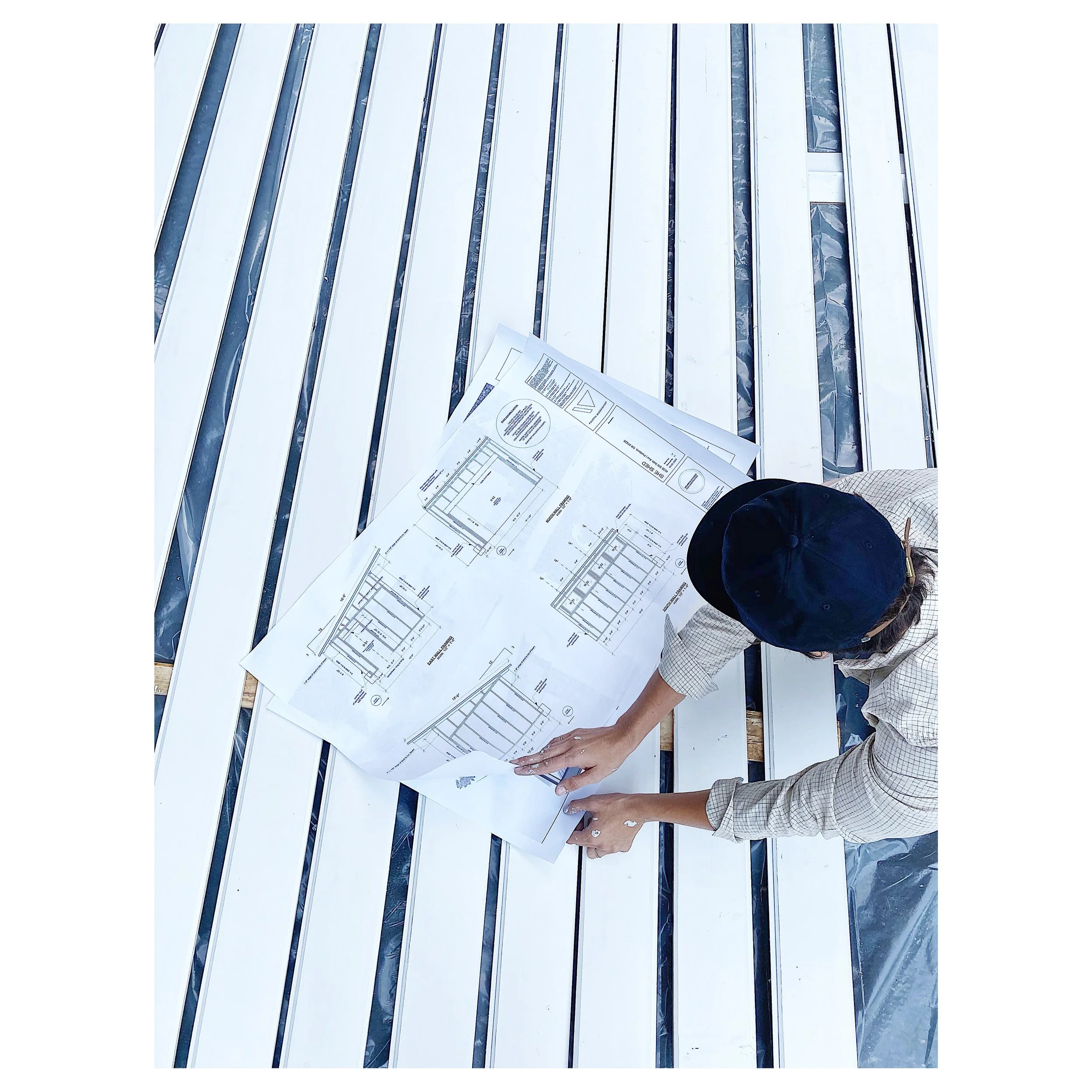 Person wearing a cap and plaid shirt laying on a white construction bench, looking at blueprints or construction plans