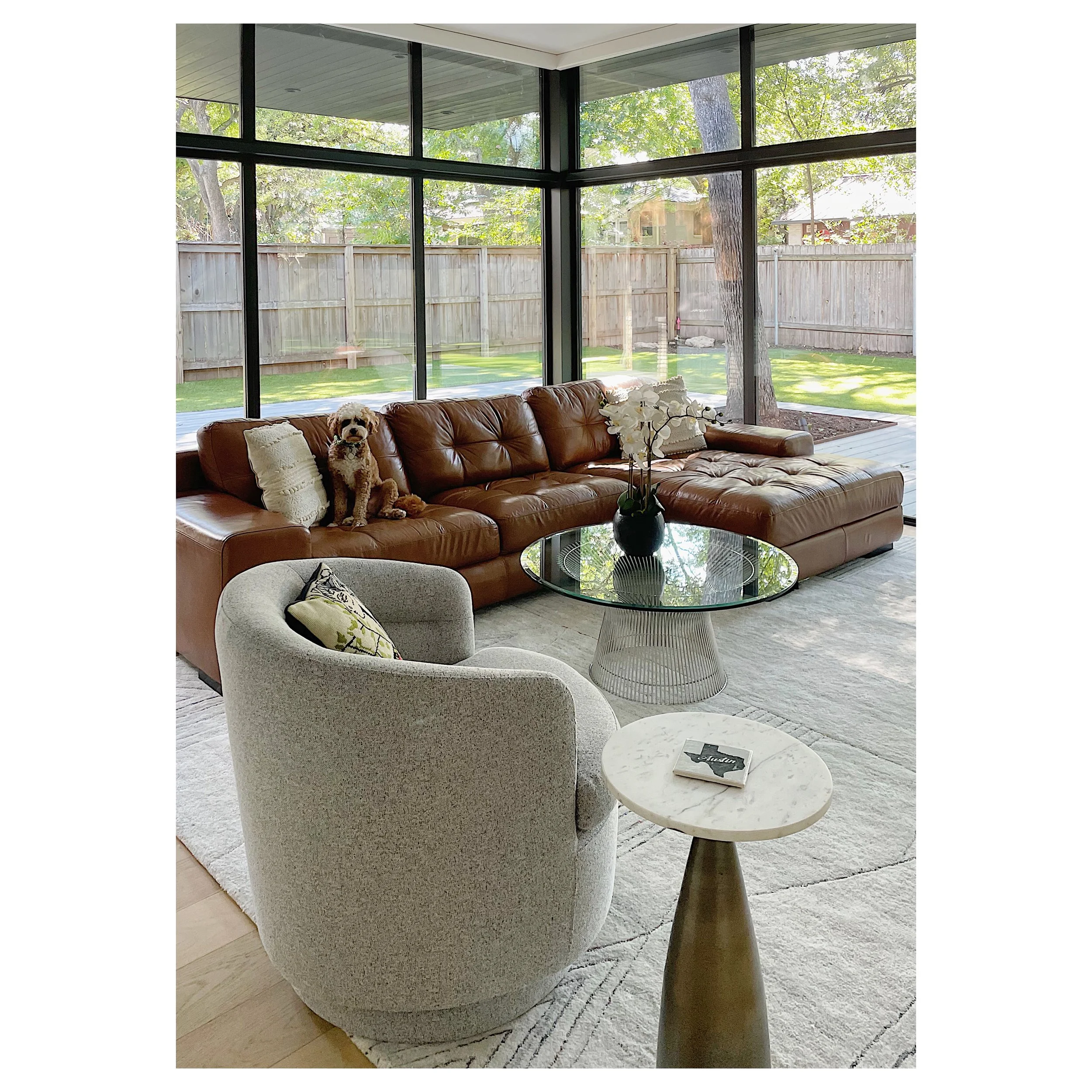 Living room with large sectional leather sofa, a white upholstered armchair, a glass coffee table with a vase of white flowers, and a small round side table with a book. Large windows show a backyard with trees and a wooden fence. A small dog sits on