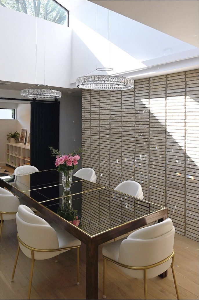 Modern dining room with a rectangular black glass-top table, surrounded by six white chairs with gold accents. A vase of pink roses is on the table, with a textured brick wall behind it. Two round chandeliers hang from the ceiling, with natural light