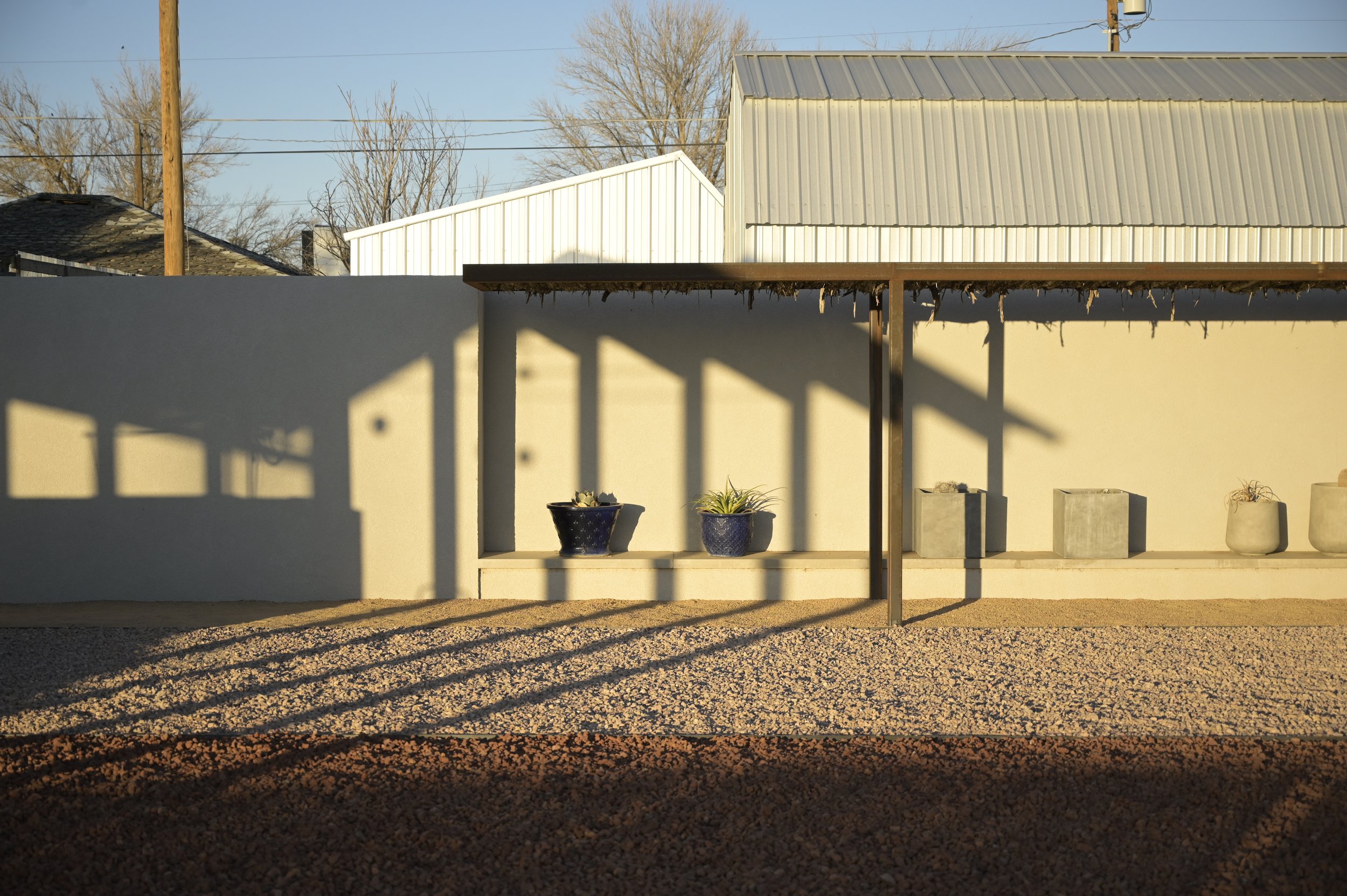 Sunlit outdoor scene with a beige wall and potted plants. Shadows cast by a structure create a pattern on the ground. Roofs and utility poles are visible in the background under a clear sky.