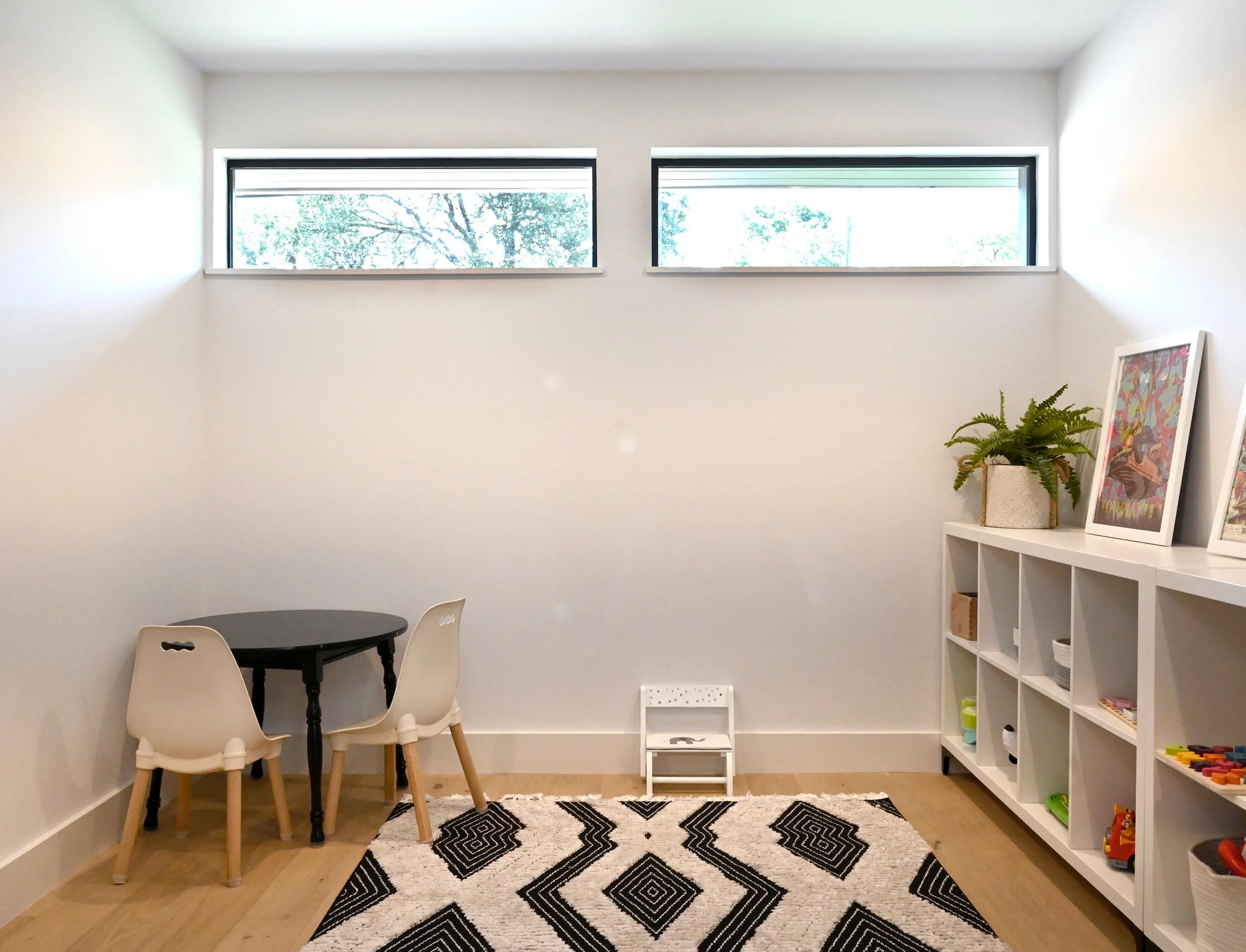 Minimalist playroom with small round black table, two white chairs, a white shelving unit with toys and framed art, a large black and white patterned rug, and a small white step stool near the wall