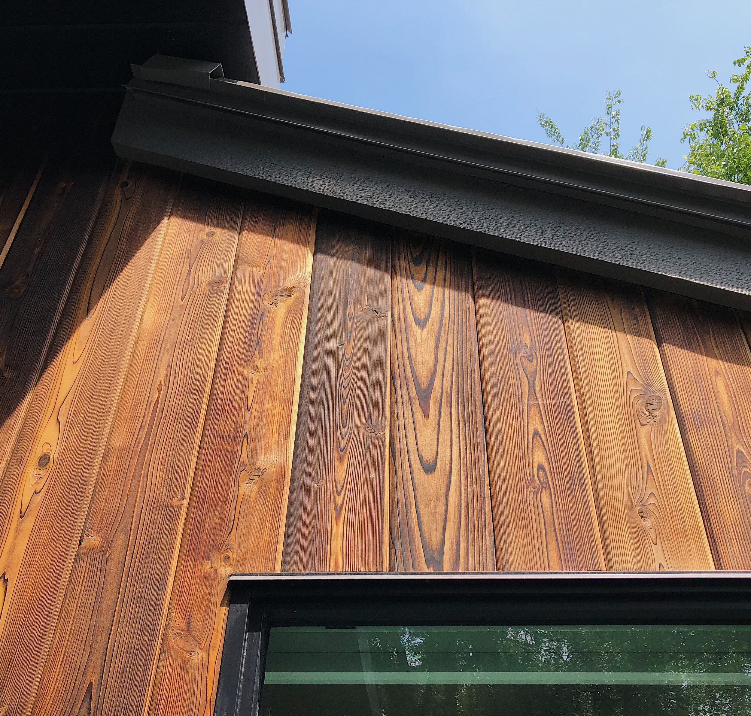 Close-up view of a wooden exterior wall with vertical planks, a black window frame, and a section of the roof with a black overhang against a blue sky with some green tree leaves.