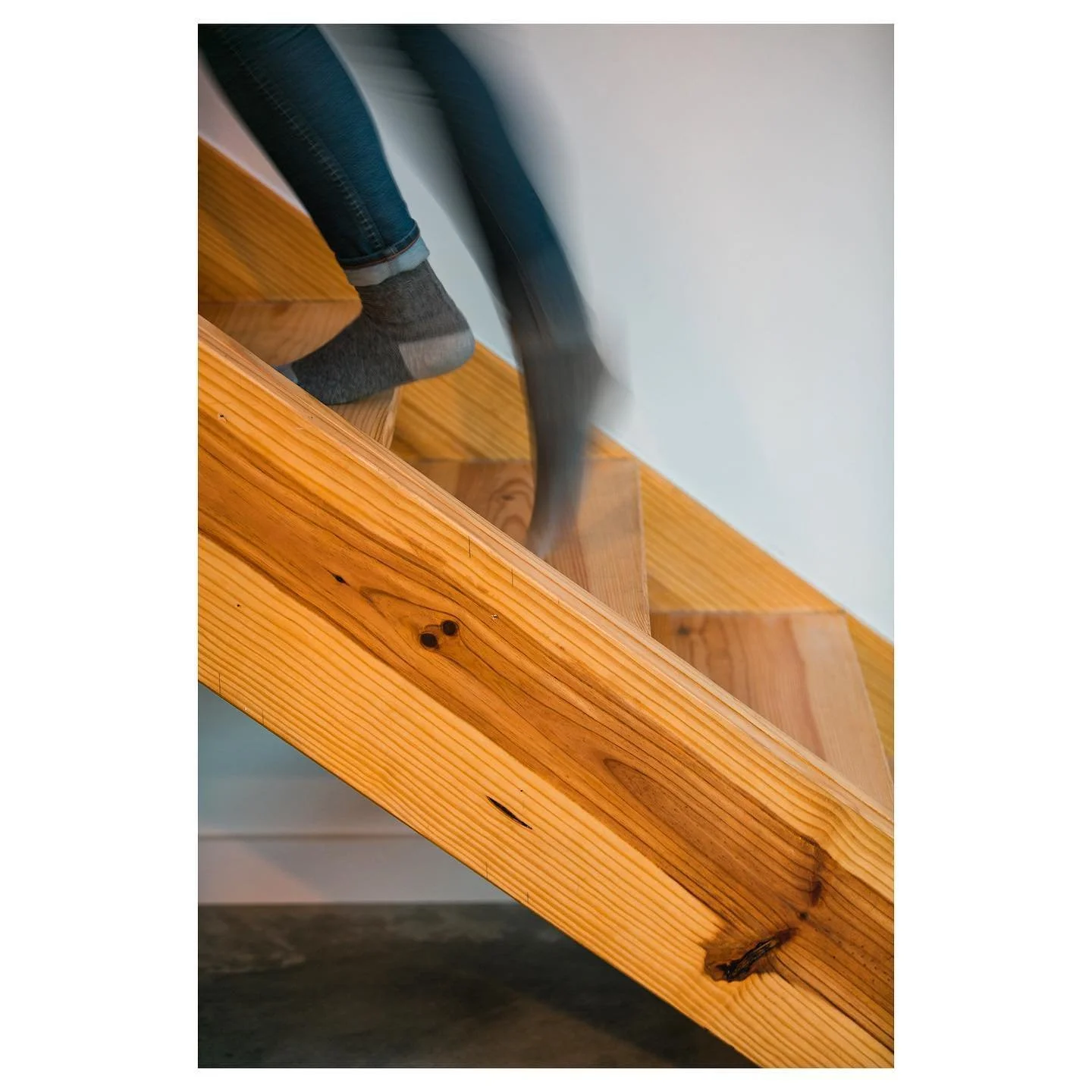 Close-up of a wooden staircase with a person's feet wearing gray socks and jeans, walking up the stairs, showing the natural wood grain and knots.