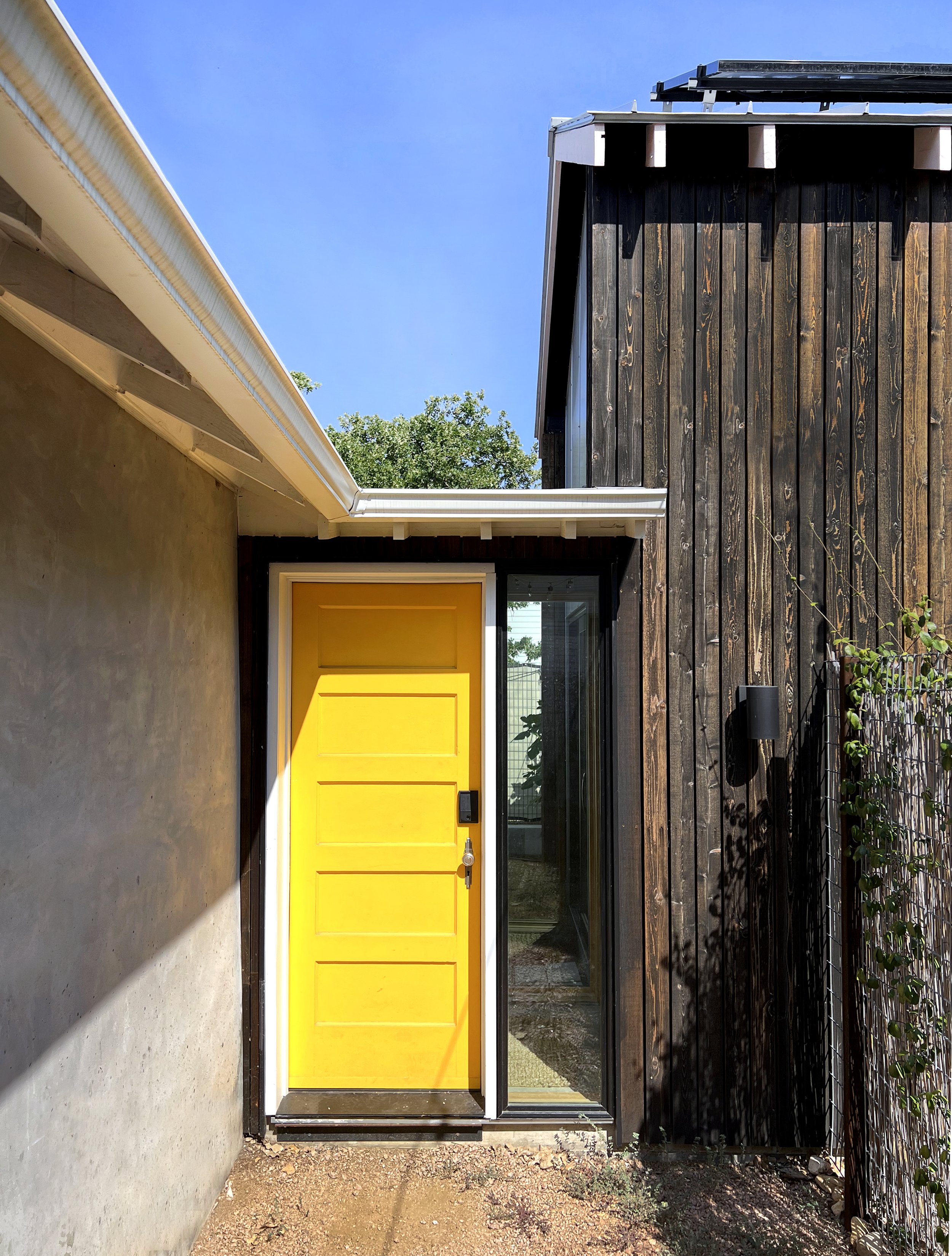 Bright yellow front door with a digital lock, beside a large window, part of a modern house with black wooden siding, a concrete wall, and a small dirt area in front, under a clear blue sky.