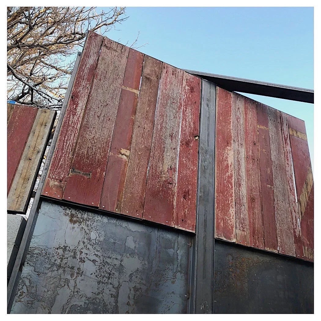 Close-up of a rusty, weathered red wooden fence attached to a metal structure with a concrete wall below, with a tree and clear blue sky in the background.