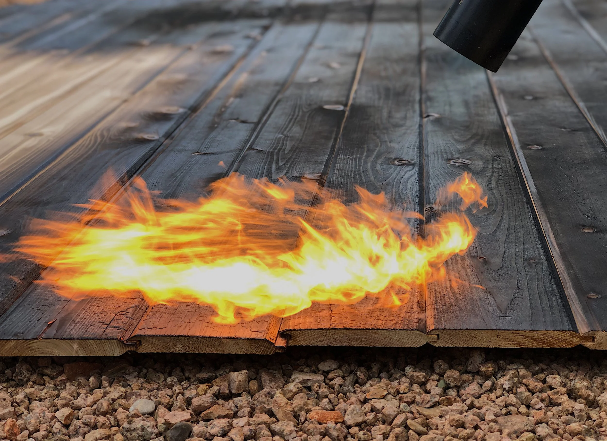 A close-up view of a fire burning on a wooden deck, with a black propane torch pointed at the flames.