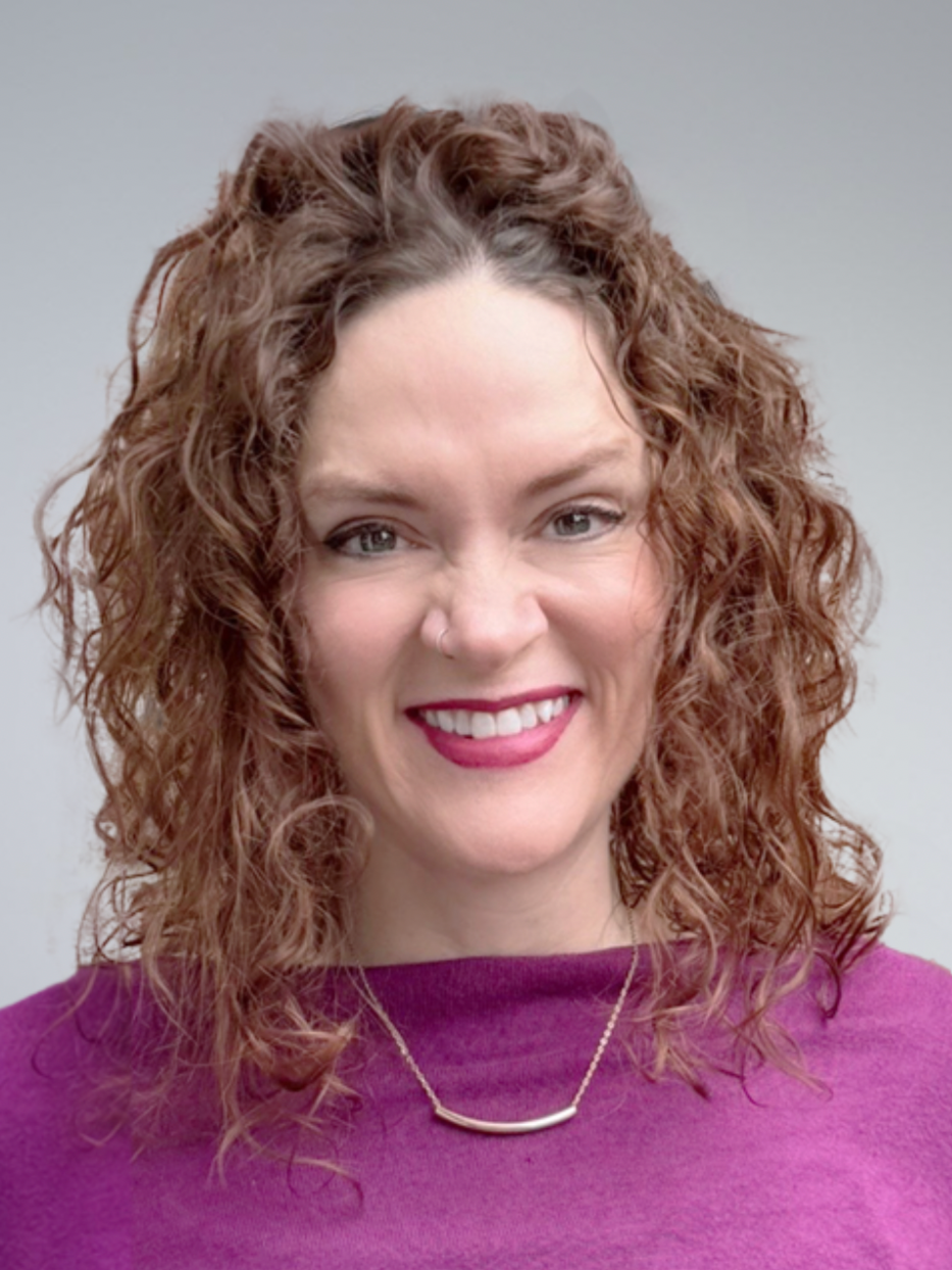 A woman with curly red hair smiling, wearing a purple top and a gold necklace, with a neutral background.