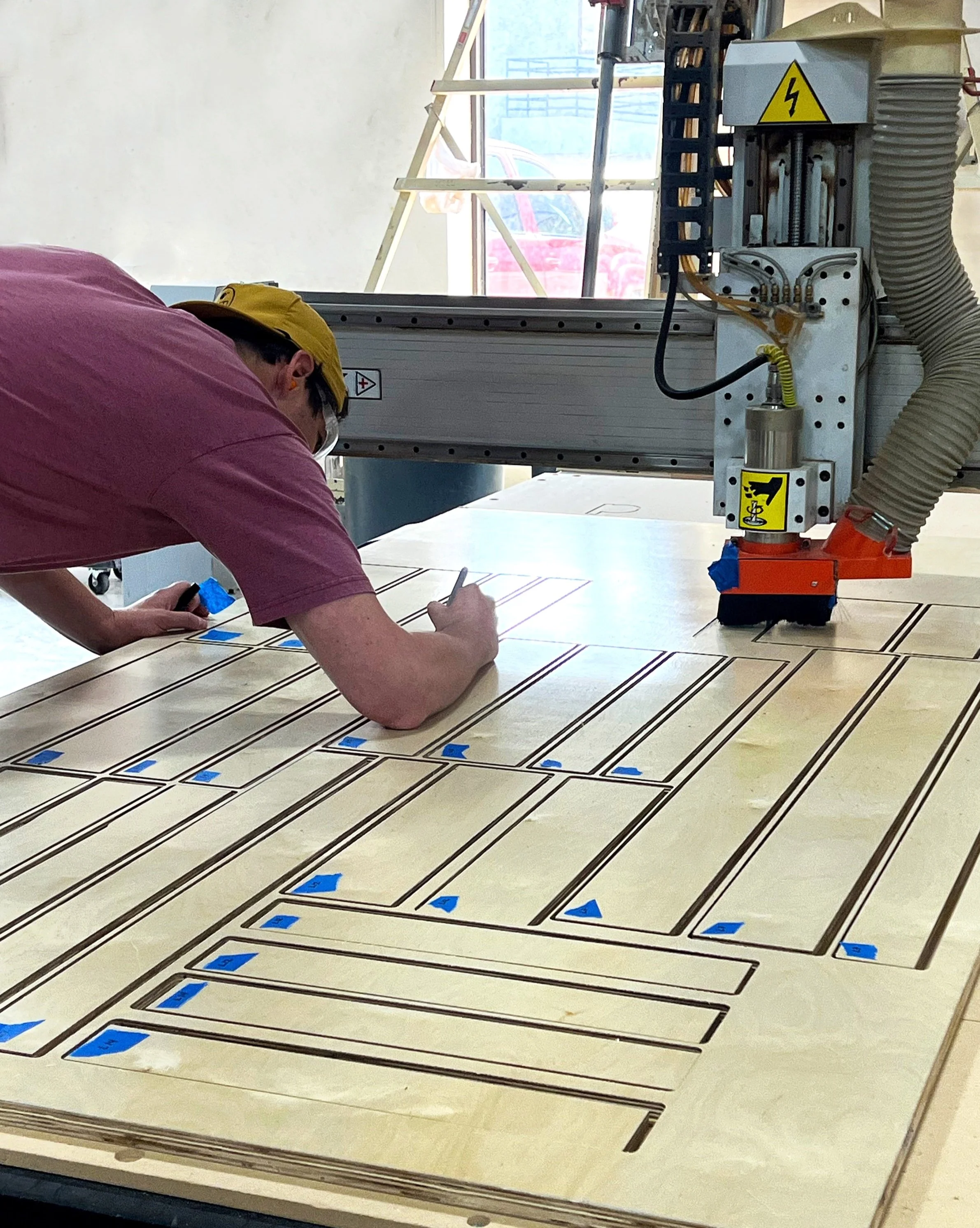 A man wearing a yellow cap and pink shirt is inspecting or working on a large wooden surface with outlined sections and blue pieces of tape. A large industrial machine with warning symbols is in the background.