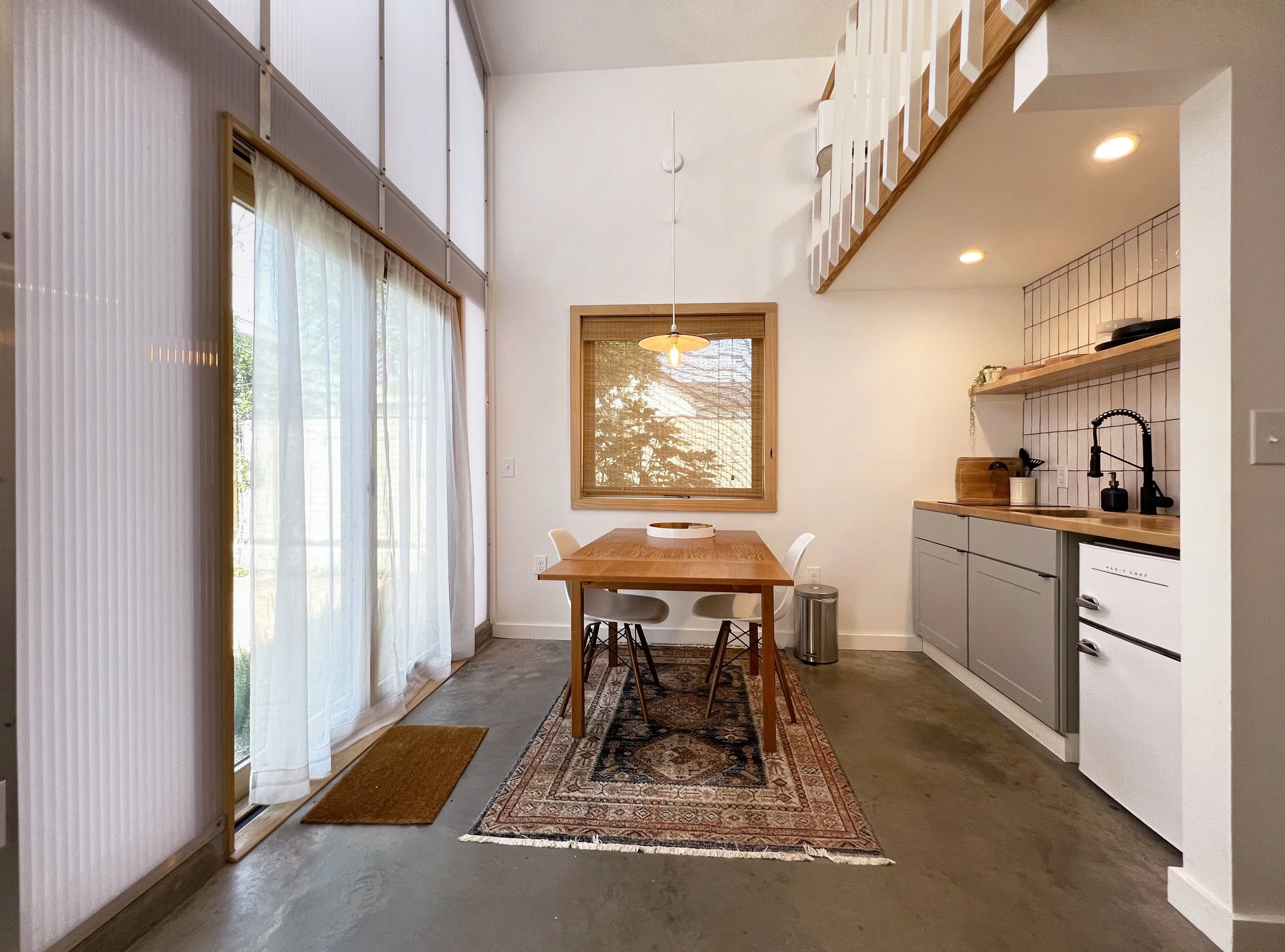 Kitchen and dining area with a wooden dining table, white chairs, a rug, and a kitchen with gray cabinets, a pink tiled backsplash, and open wooden shelves.