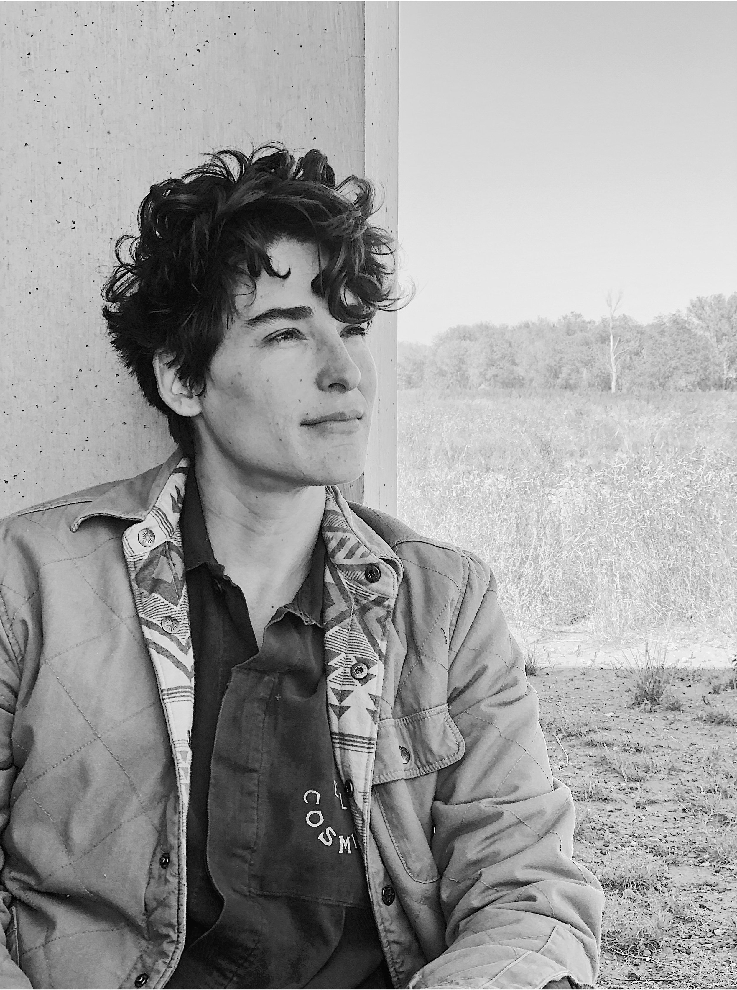 Black and white photo of a young woman with short curly hair, sitting outdoors near a concrete wall, gazing into the distance with a contemplative expression.