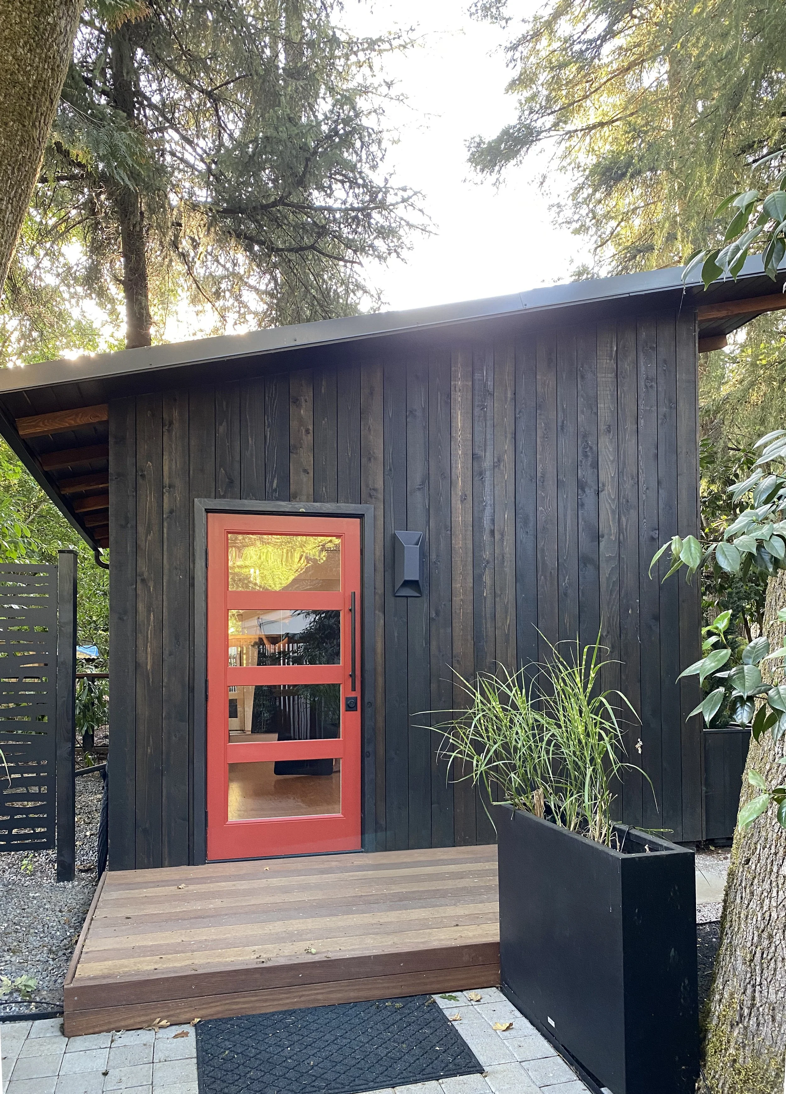 A small modern black wooden building with a red door, a deck in front, and closed black mailbox or speaker on the wall. Green trees surround it.