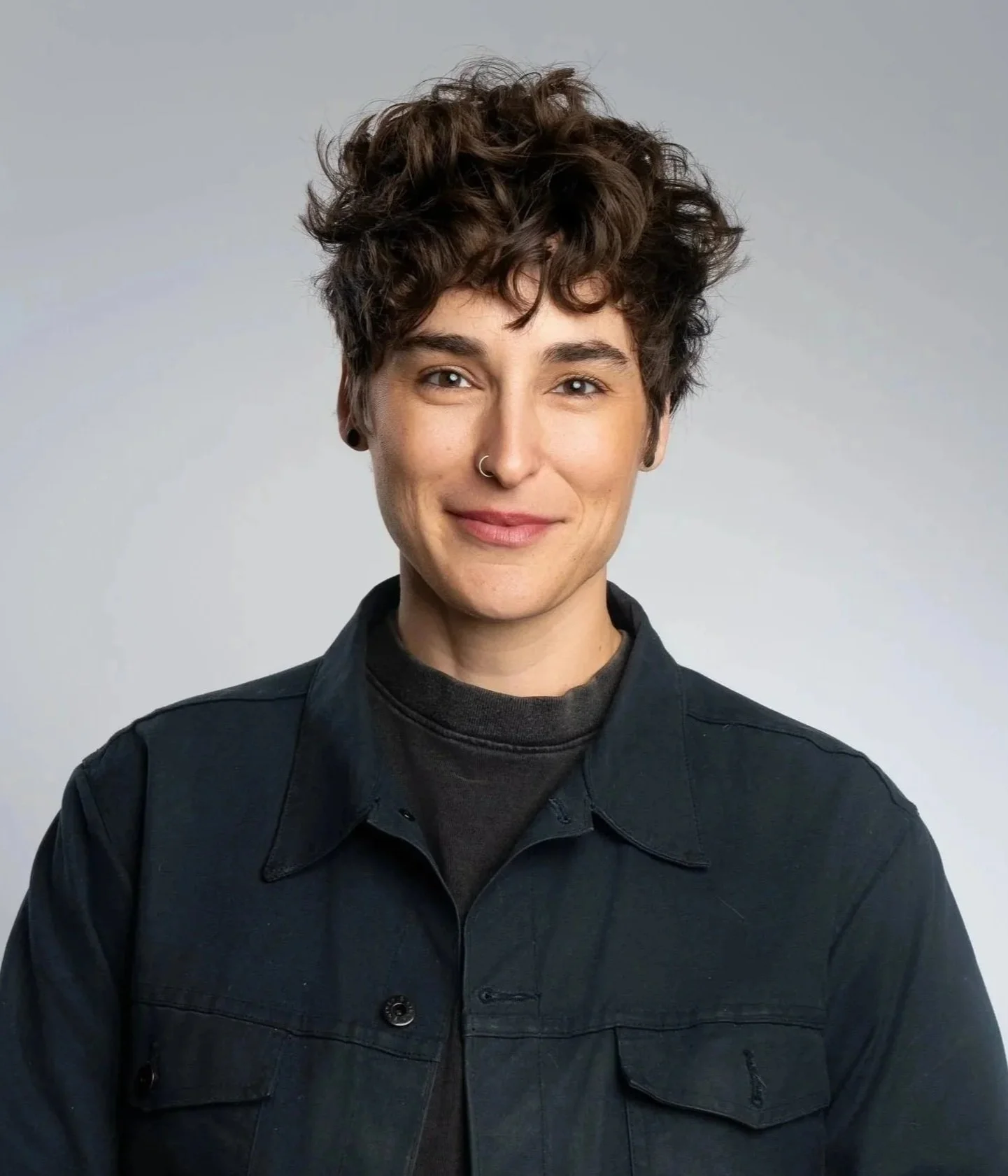 A woman with short curly brown hair, nose piercing, and black earrings, wearing a black jacket and dark shirt, smiling in front of a light gray background.
