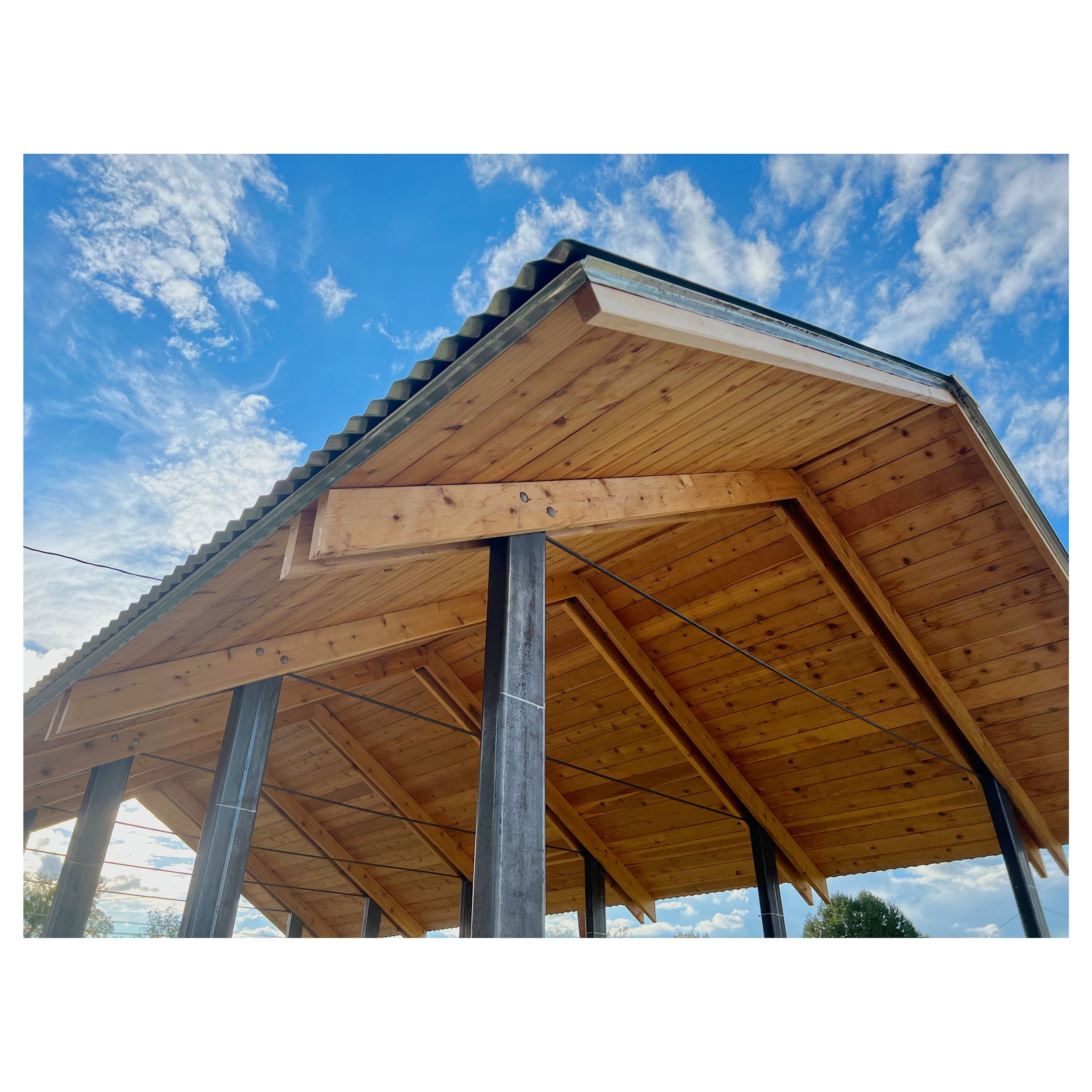 Wooden building under construction with steel supports, a metal roof, and a blue sky with scattered clouds in the background.