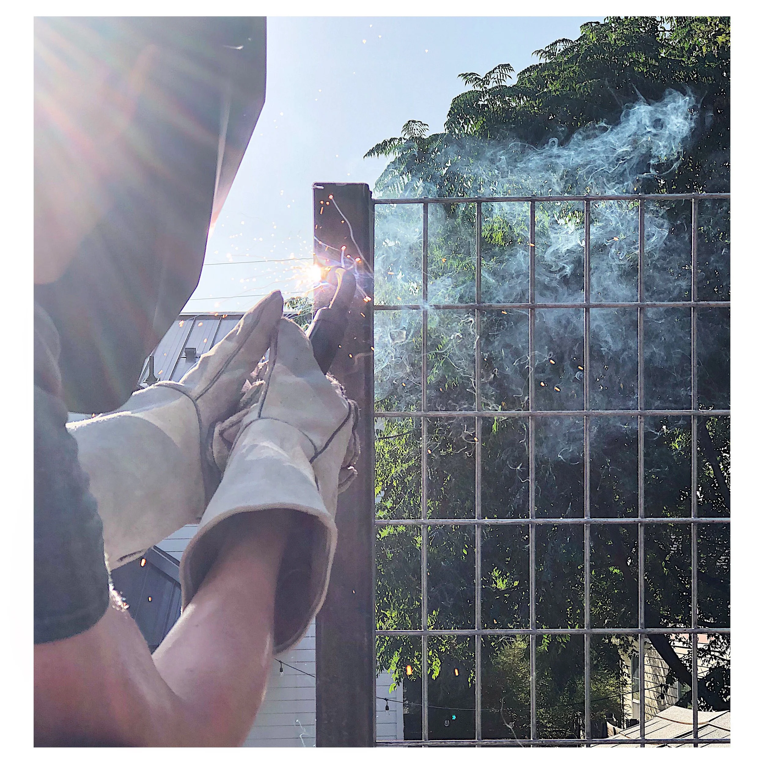 Person welding on a metal fence outdoors, sparks and smoke visible, with sunlight shining in the background.