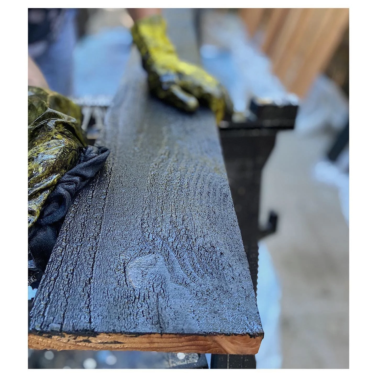 Close-up of a wooden surface being painted with black stain by a person wearing yellow and black gloves.