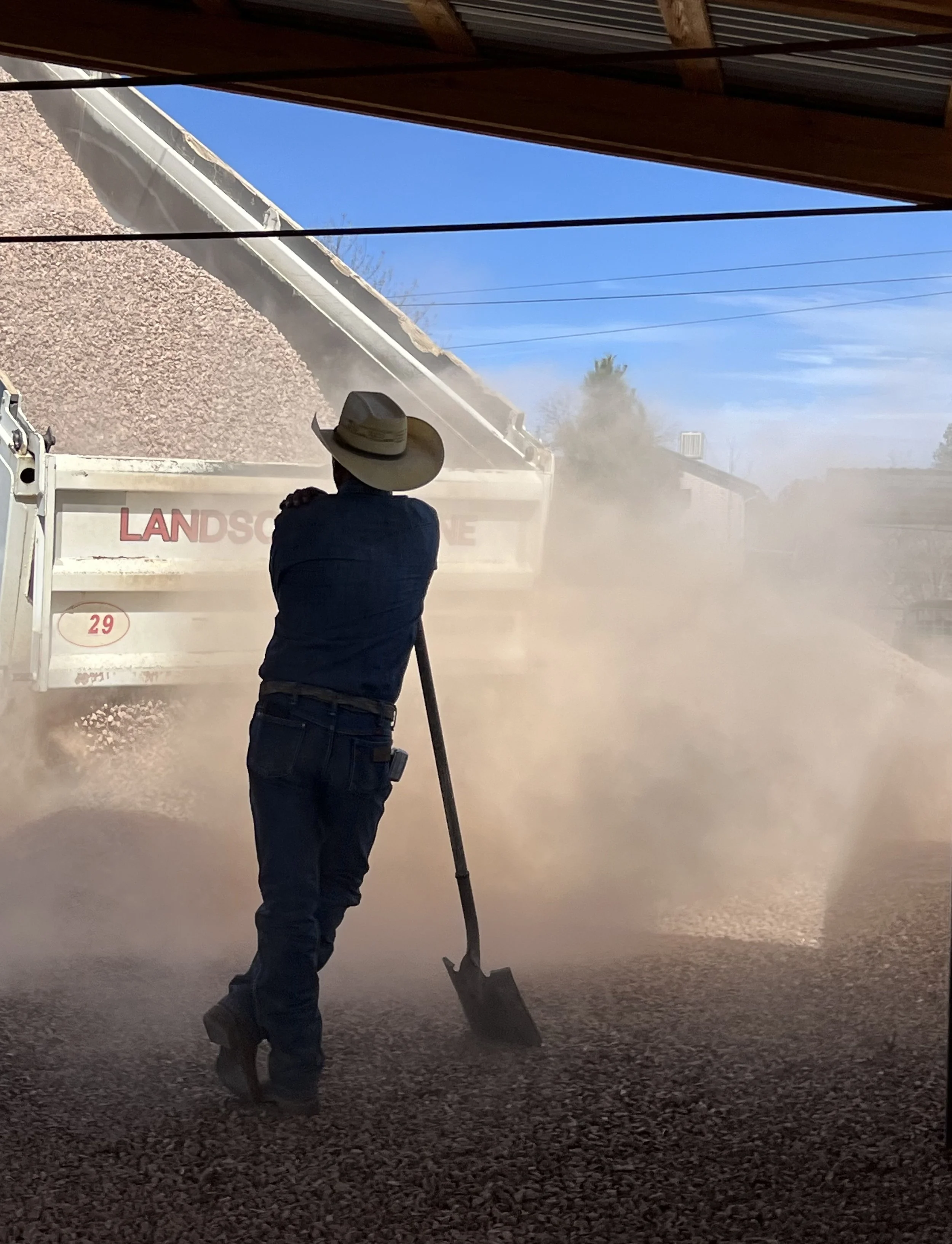 Person wearing a cowboy hat and jeans shoveling gravel during daytime with a dust cloud, a dump truck labeled 'LANDSCAPE' in the background, clear blue sky, and overhead power lines.
