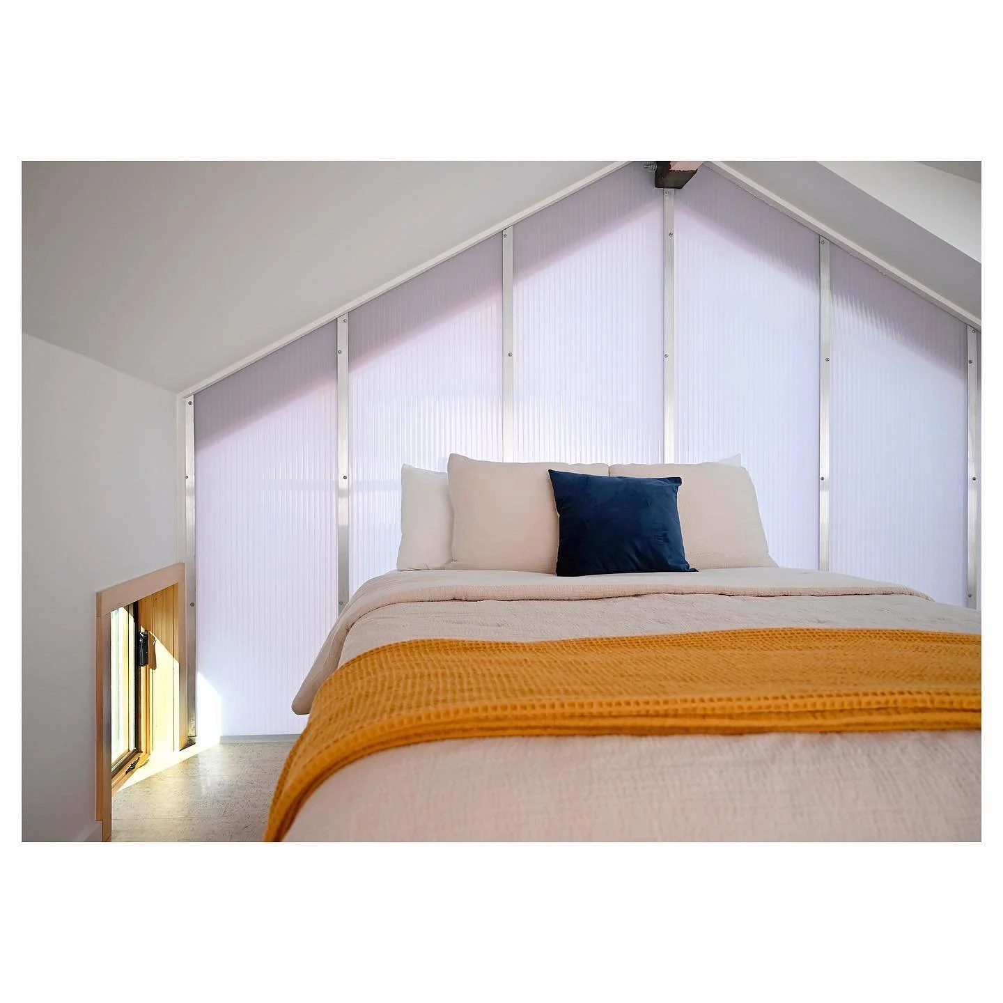 A cozy bedroom with a sloped ceiling, featuring a neatly made bed with white and beige bedding, a dark blue pillow, and an orange throw blanket. There is a small window on the left side, allowing natural light into the room.