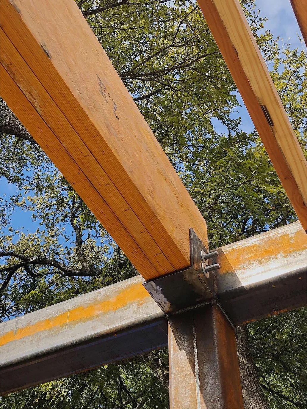 Close-up view of a wooden construction beam attached to a metal support structure, with green trees and a blue sky in the background.