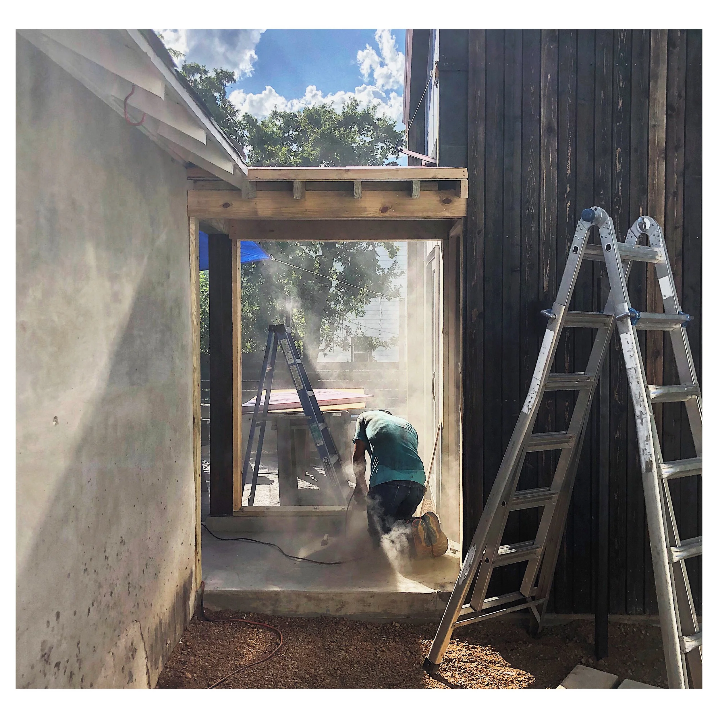 Worker kneeling and working on a wooden doorway frame under sunny skies, with ladders and construction dust visible.