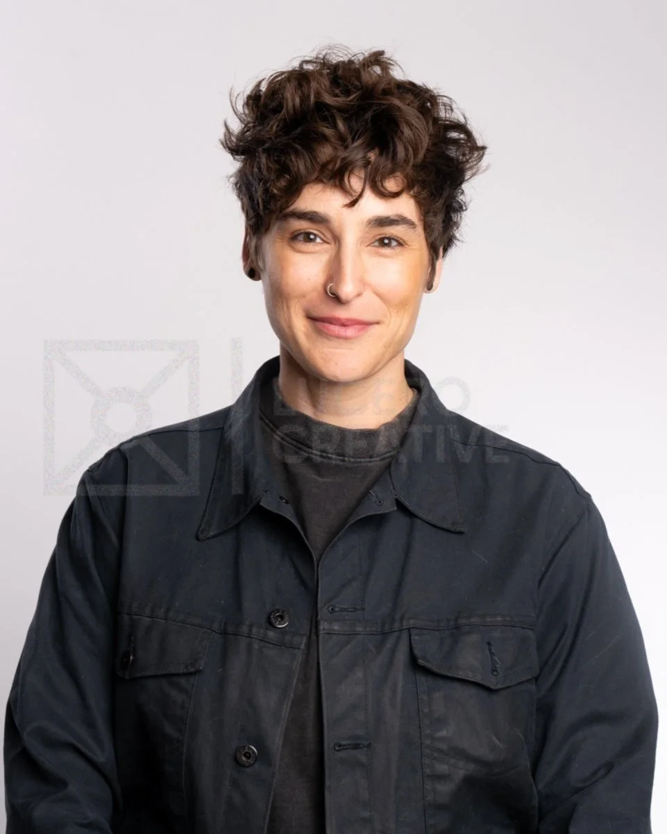 A person with short, curly brown hair, wearing a black jacket and a dark shirt, smiling at the camera.