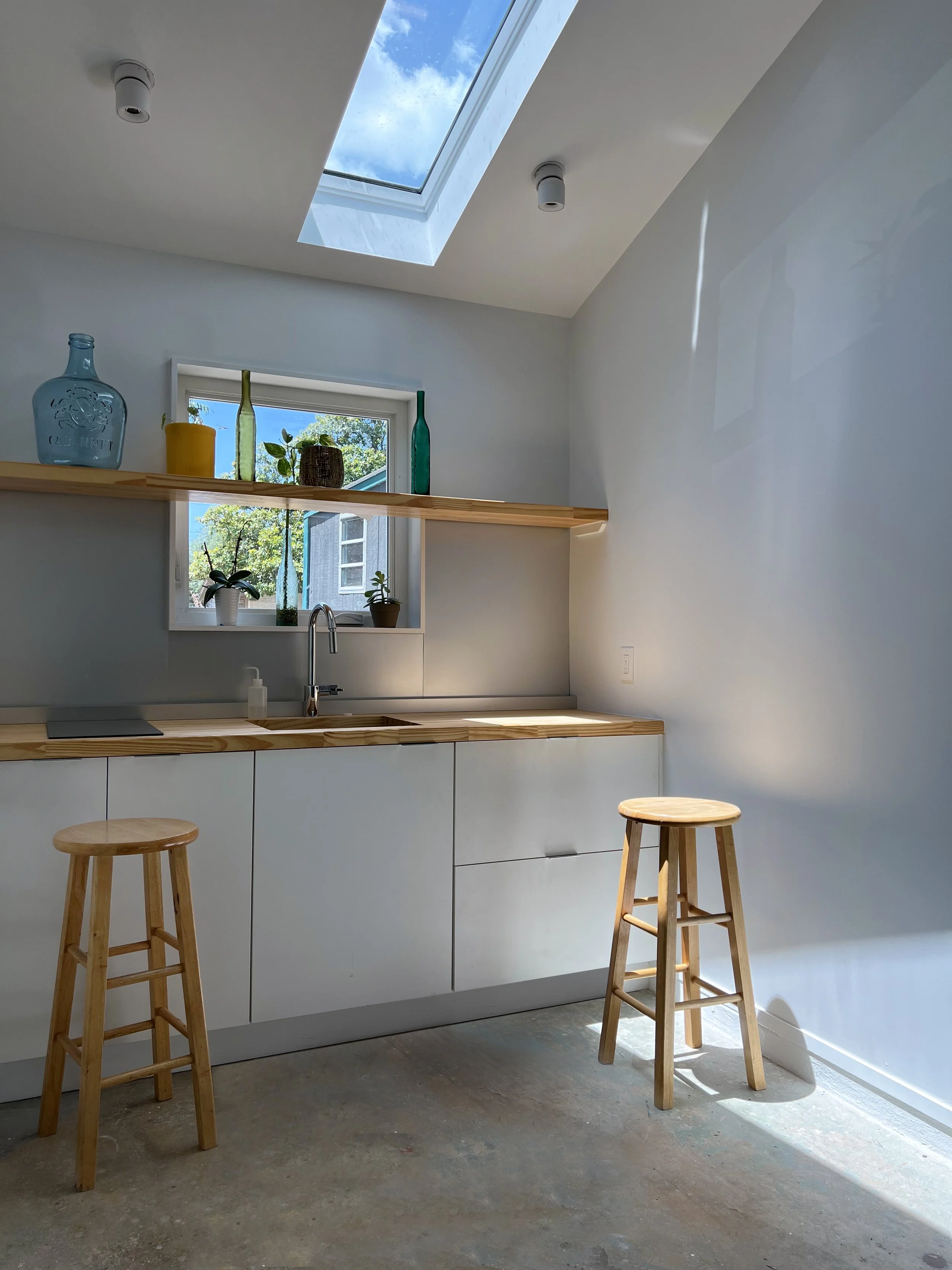 Minimalist kitchen with white cabinets, wooden countertops, two wooden stools, a window with potted plants, and a skylight letting in natural light.