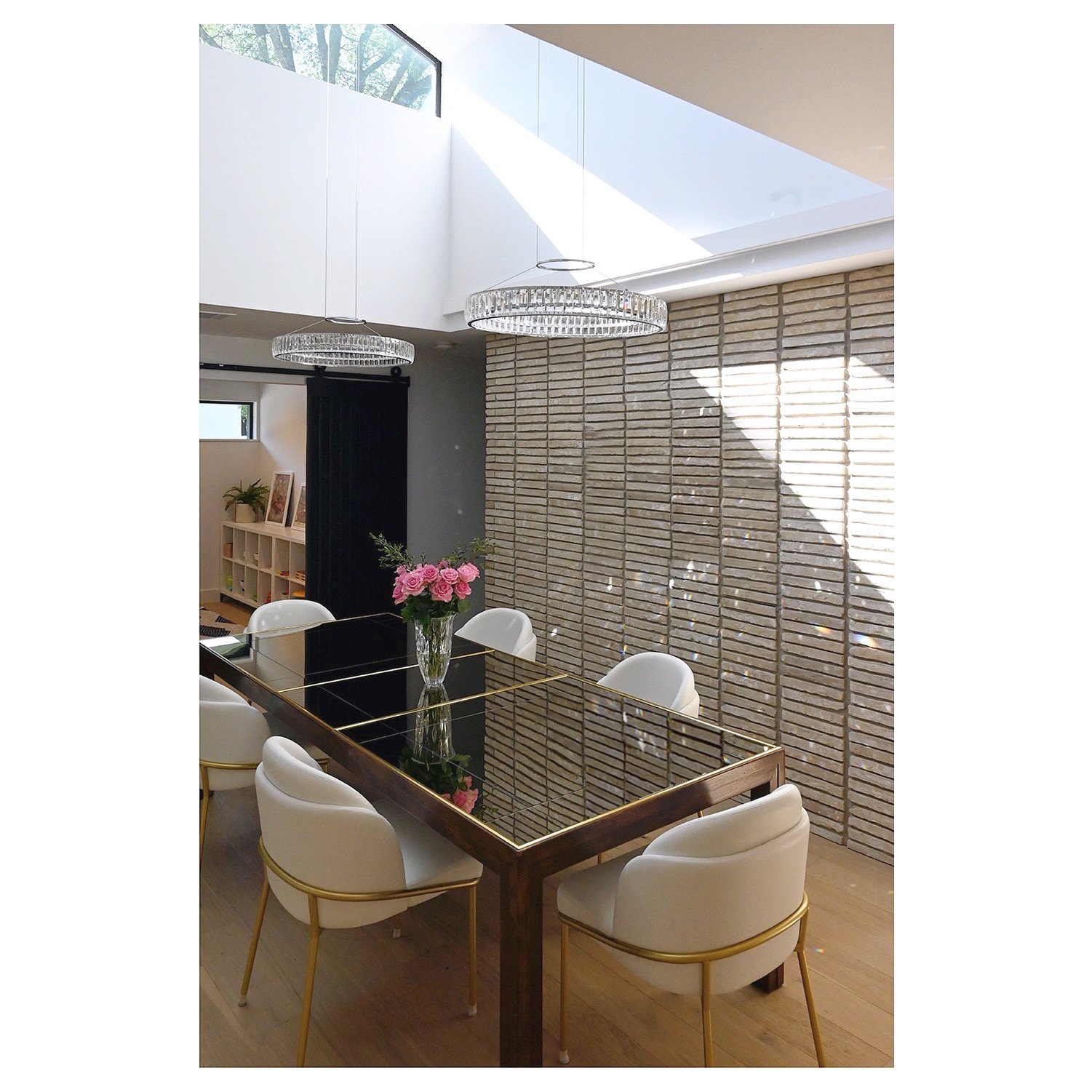 A modern dining room featuring a glass-topped wooden table with gold accents, surrounded by six white upholstered chairs with gold legs. A vase with pink roses sits at the center of the table. The room has a brick wall and large skylight windows lett