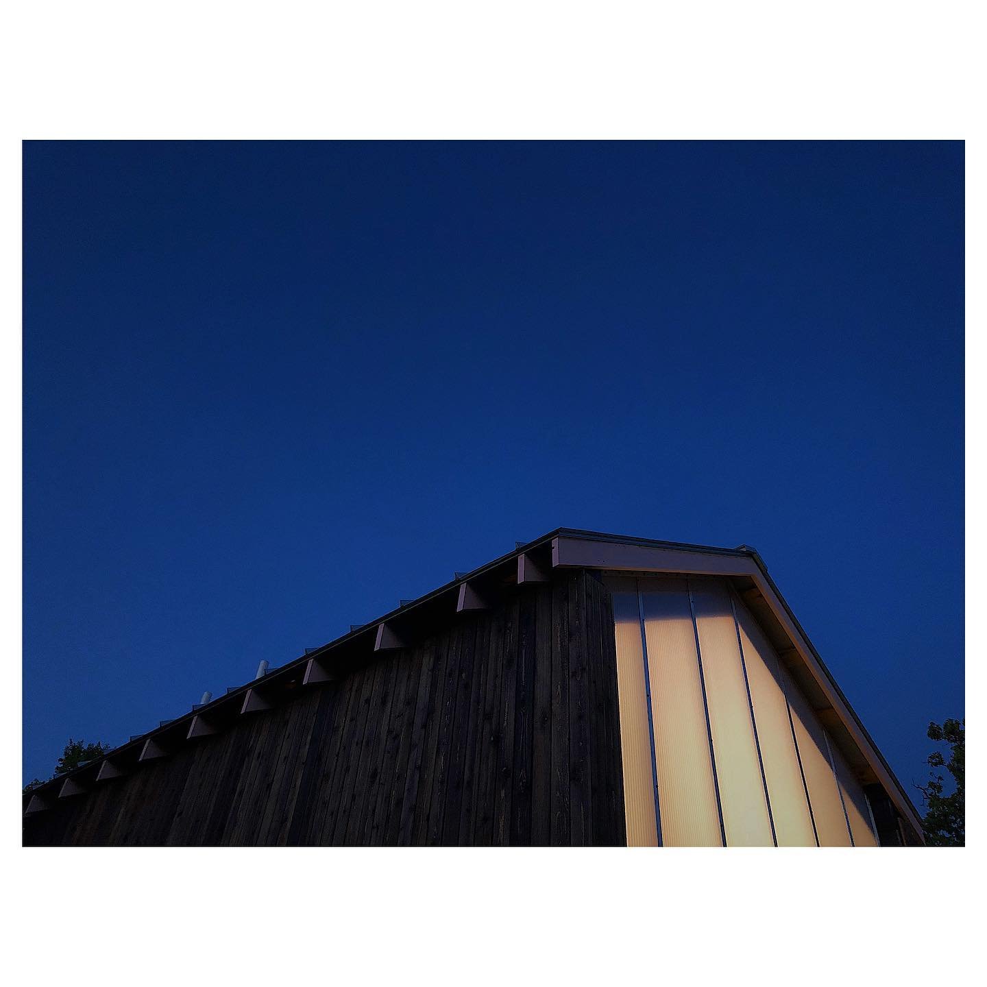 A modern building with dark wooden exterior walls and a sloped roof illuminated by indoor lights, against a dark evening sky.