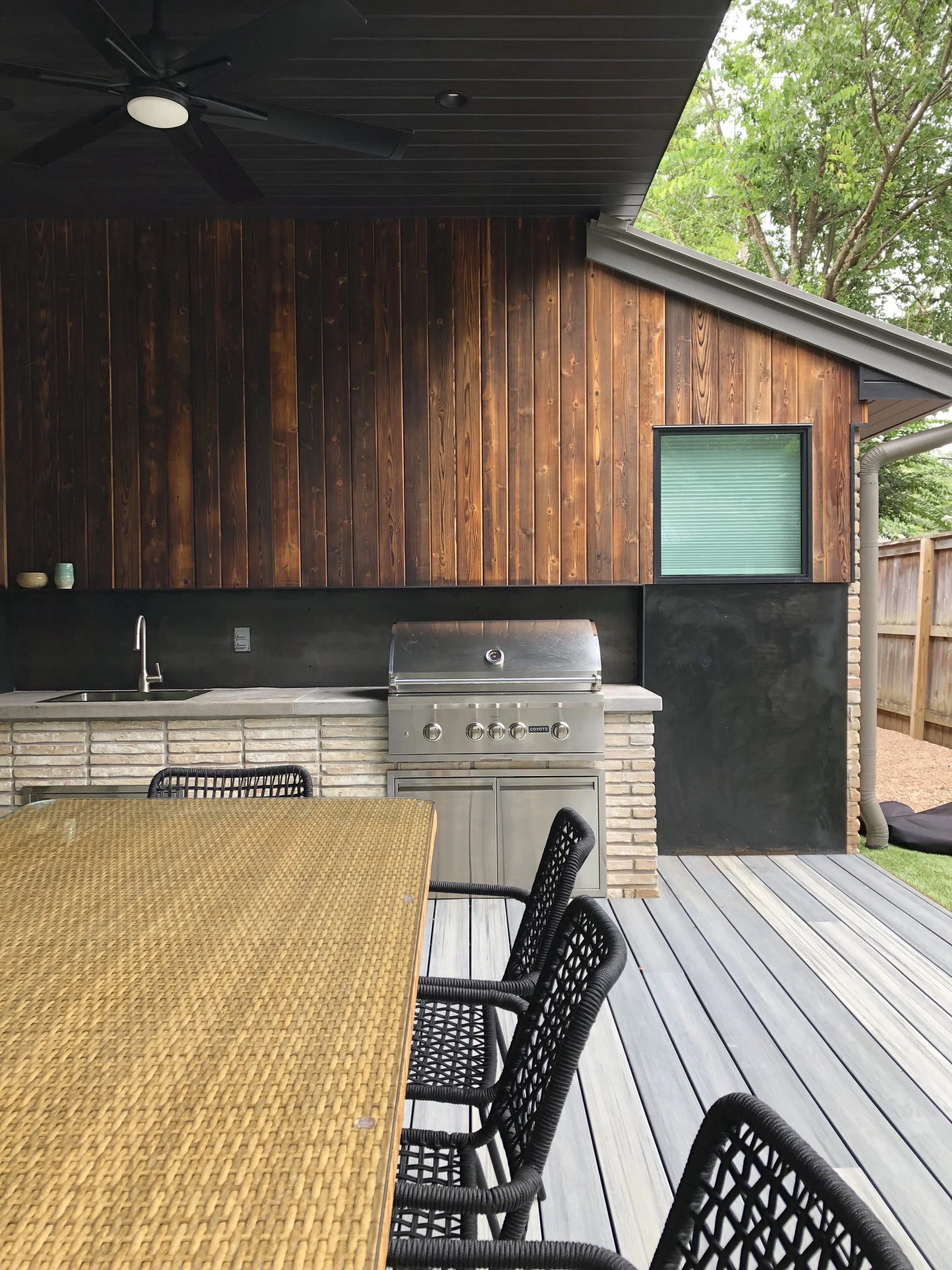 Outdoor patio with a wooden-paneled wall, a stainless steel grill, a table with woven chairs, and a ceiling fan under a covered area, with trees in the background.
