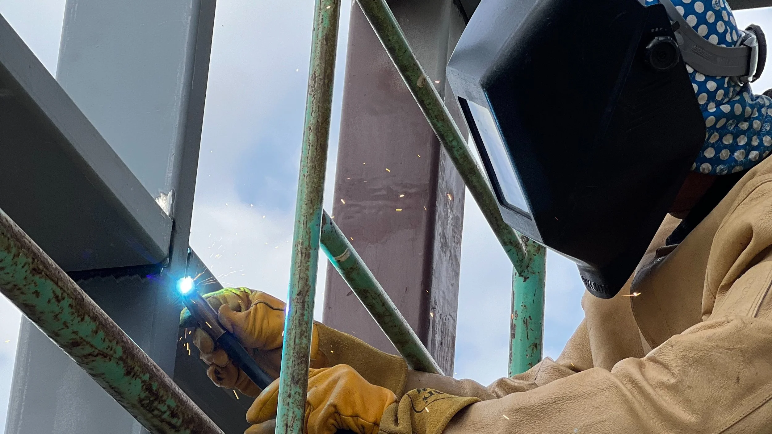 Close-up of a worker welding metal on a scaffold, wearing protective gloves and jacket, with sparks flying. The worker's face is not visible.