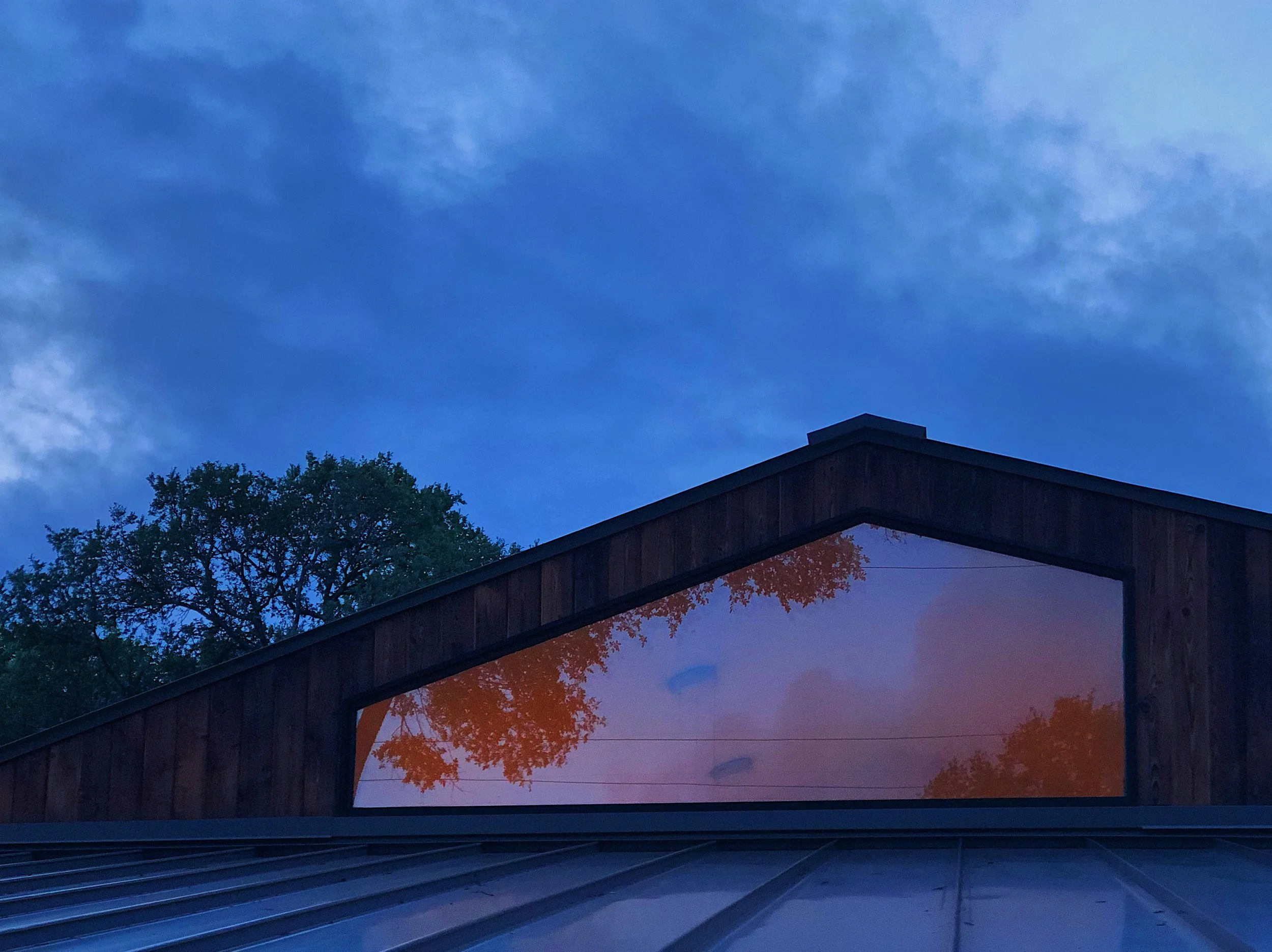 Reflection of orange autumn leaves and blue sky in a triangular window on a wooden building's sloped roof at dusk.
