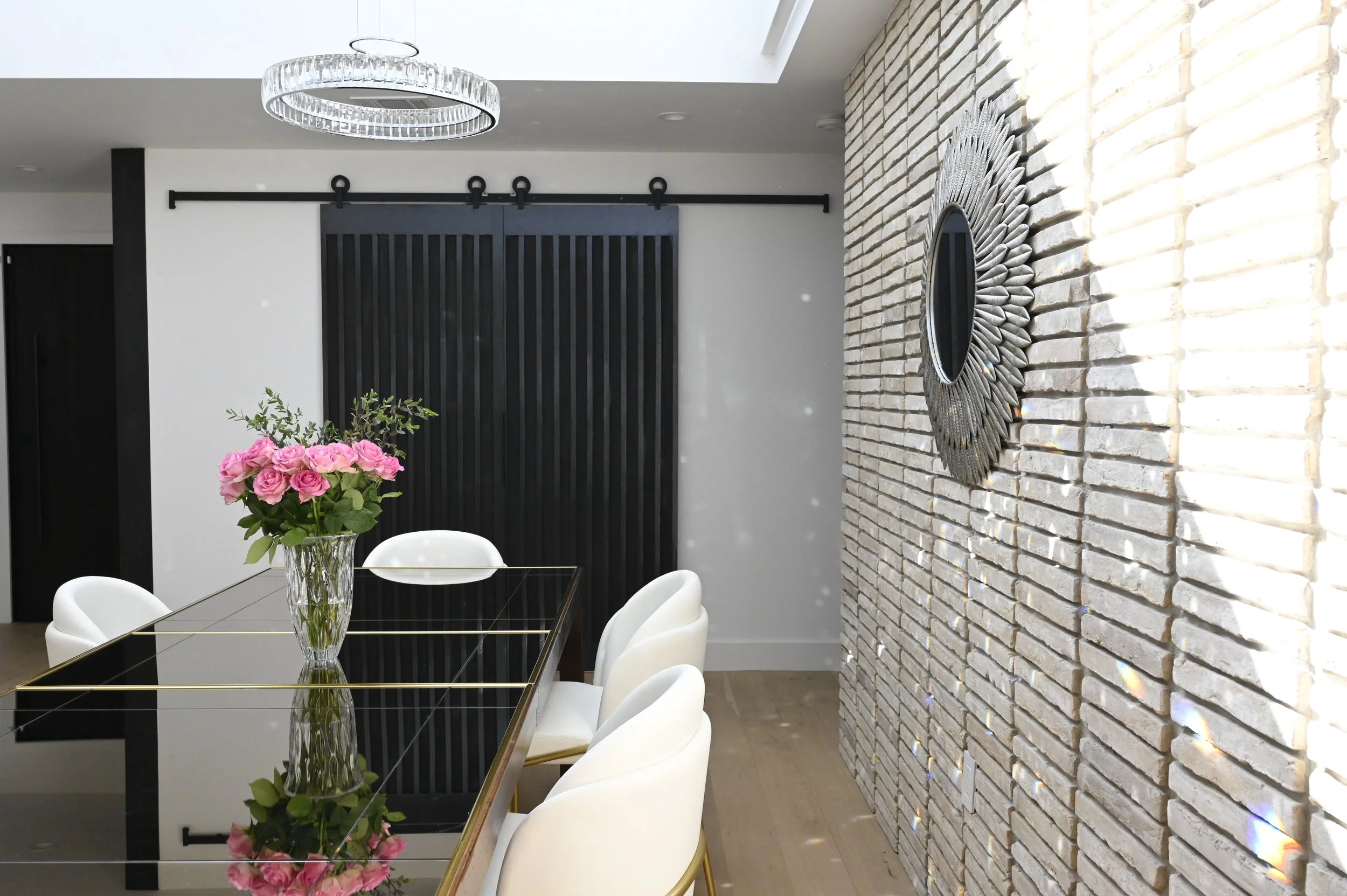 A modern dining room with a glass table, white chairs, a vase of pink roses, a black sliding barn door, a circular crystal chandelier, and a decorative circular mirror on a white brick wall.