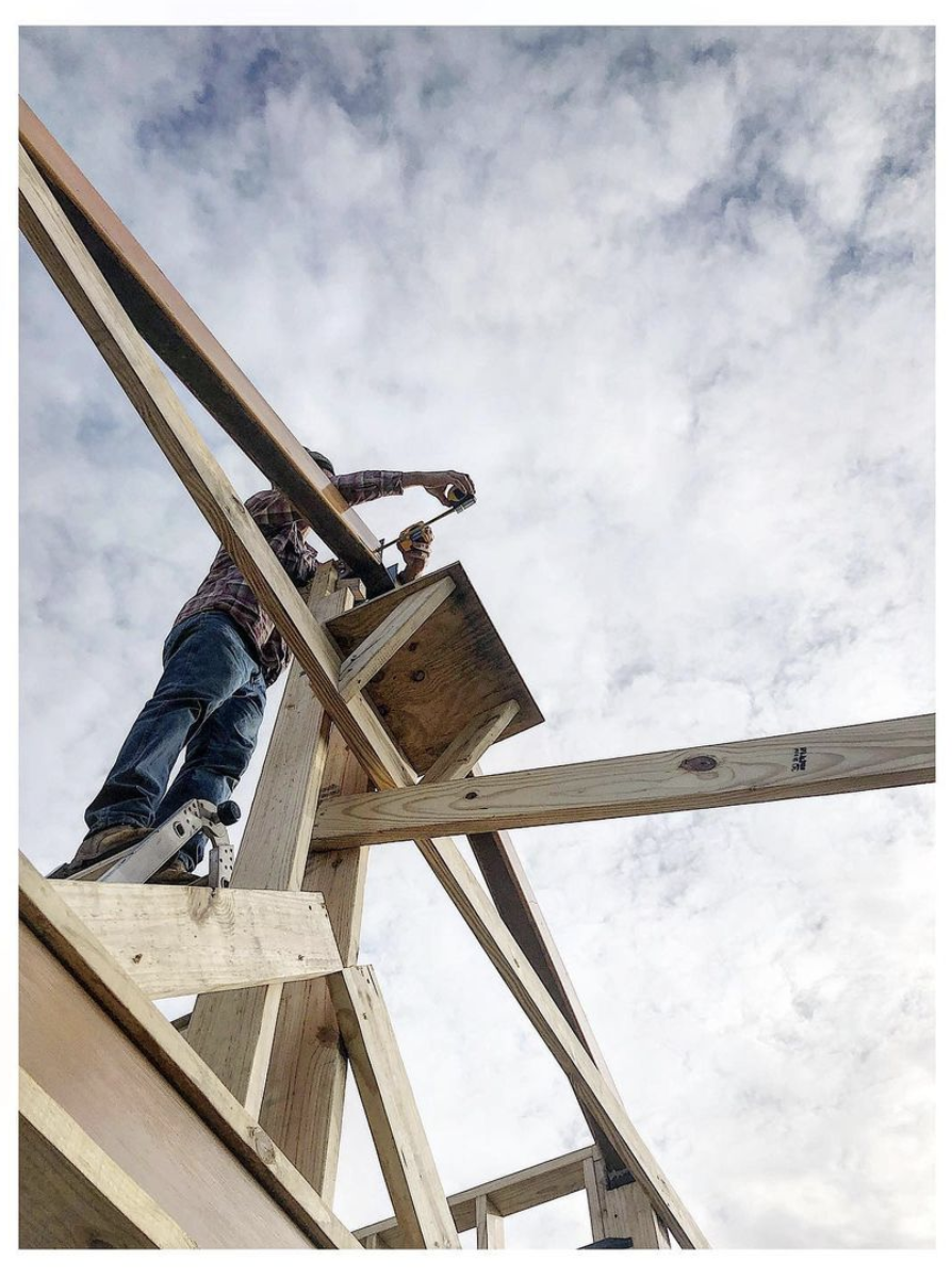 A person working on a wooden construction project, standing on a ladder and fastening a nail into a beam outdoors against a cloudy sky.