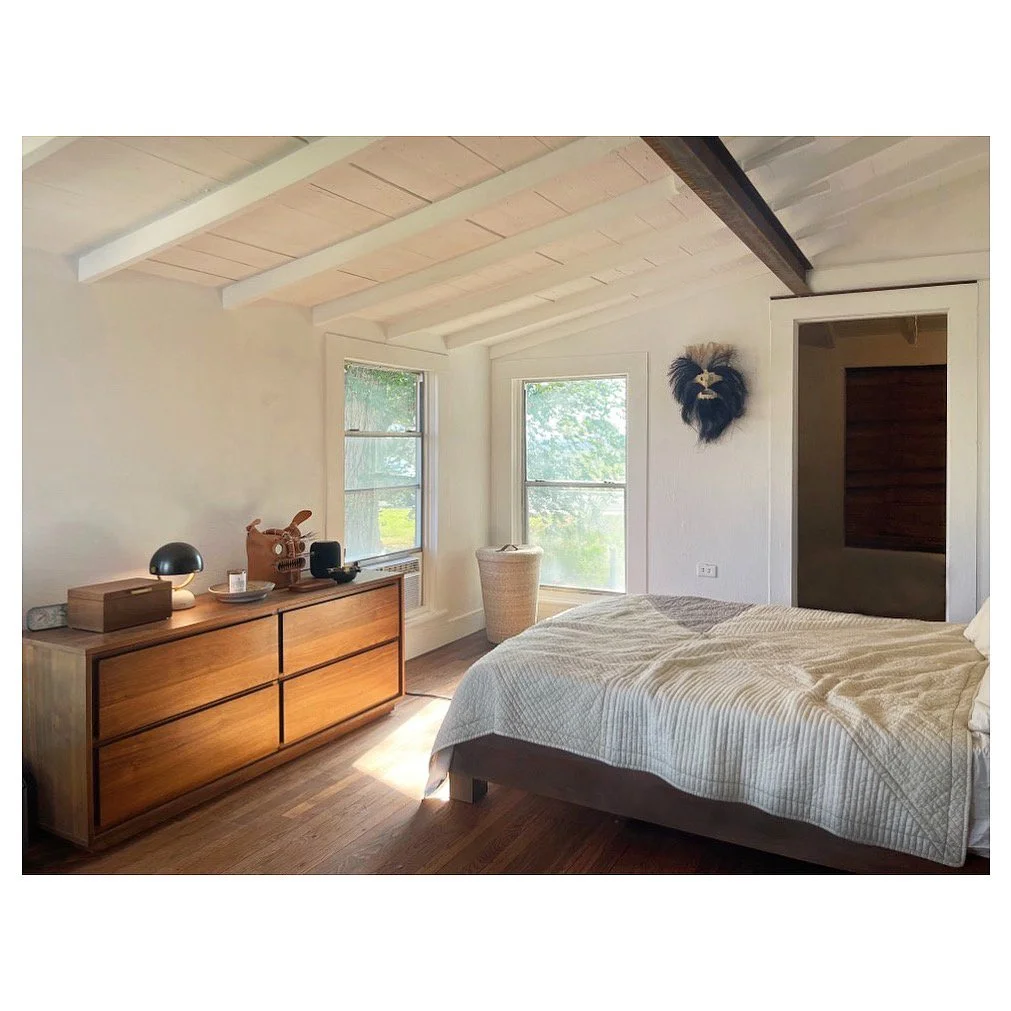Bedroom with wooden dresser, bed with beige quilt, two windows showing greenery, wall decor resembling a lion's face, wicker basket by window, and exposed ceiling beams.
