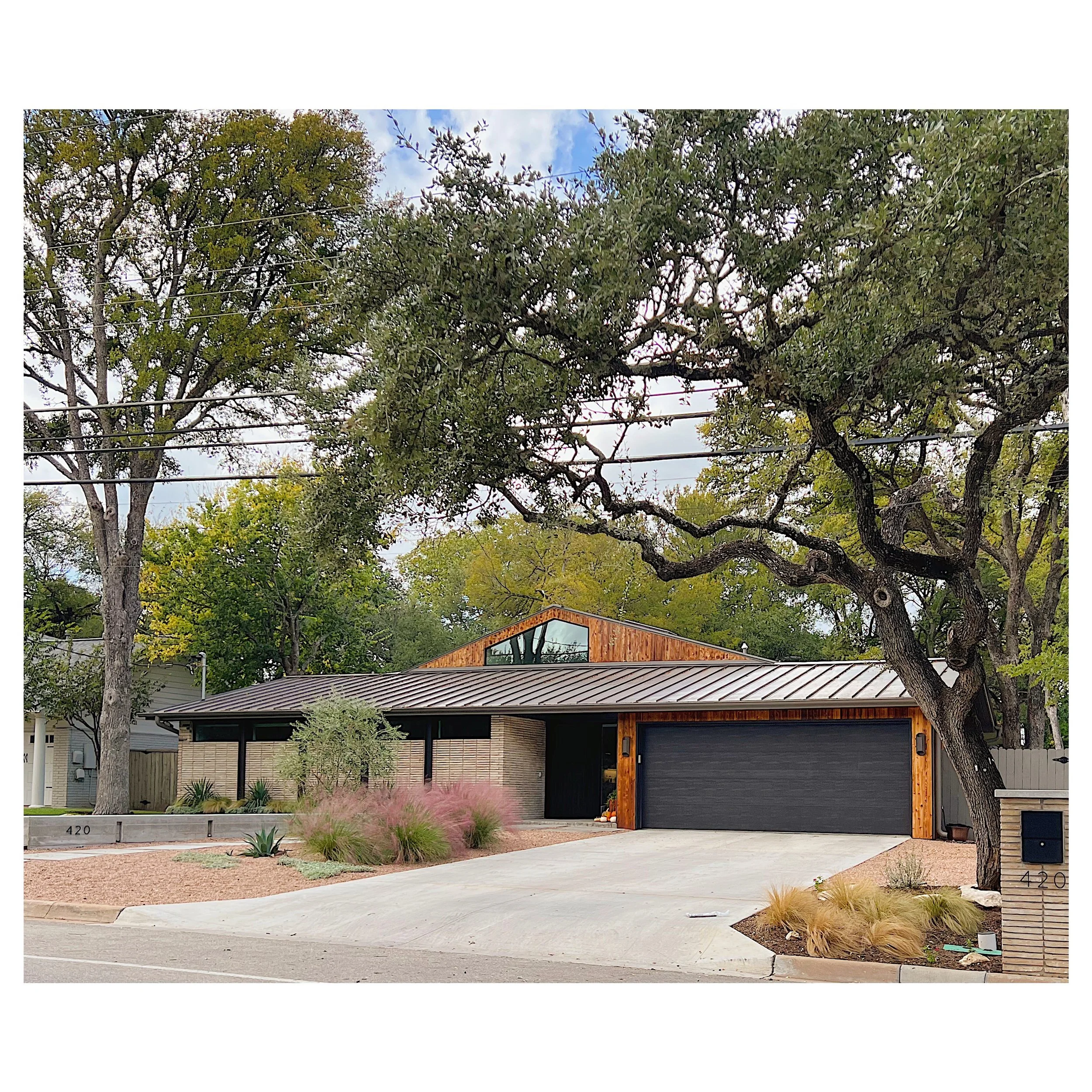 Modern house with a black garage door and wooden trim, surrounded by trees and desert landscaping, with a driveway and house numbers 420