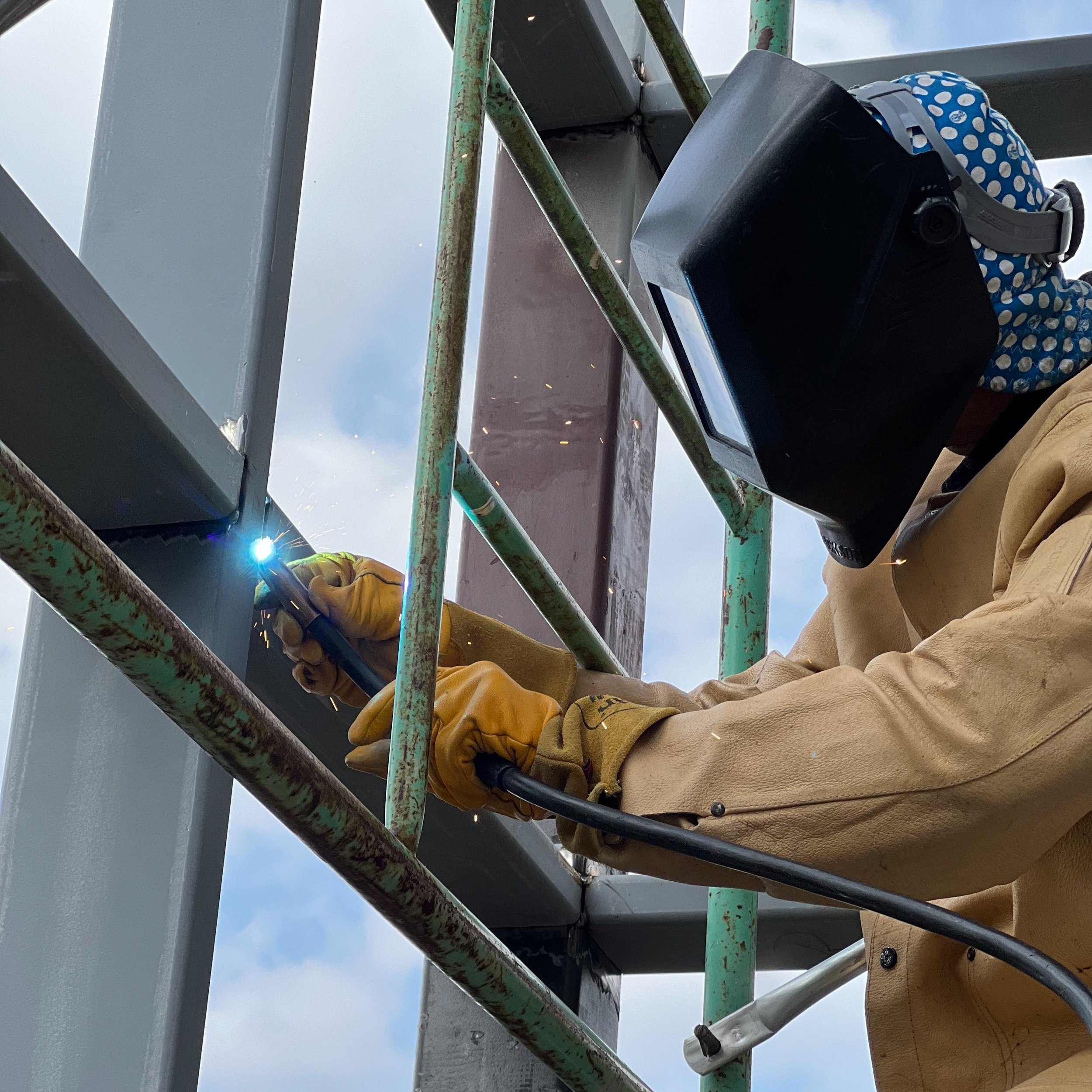 Welding a metal structure, person wearing a welding helmet, yellow gloves, and beige jacket, working outdoors with cloudy sky and green scaffolding.