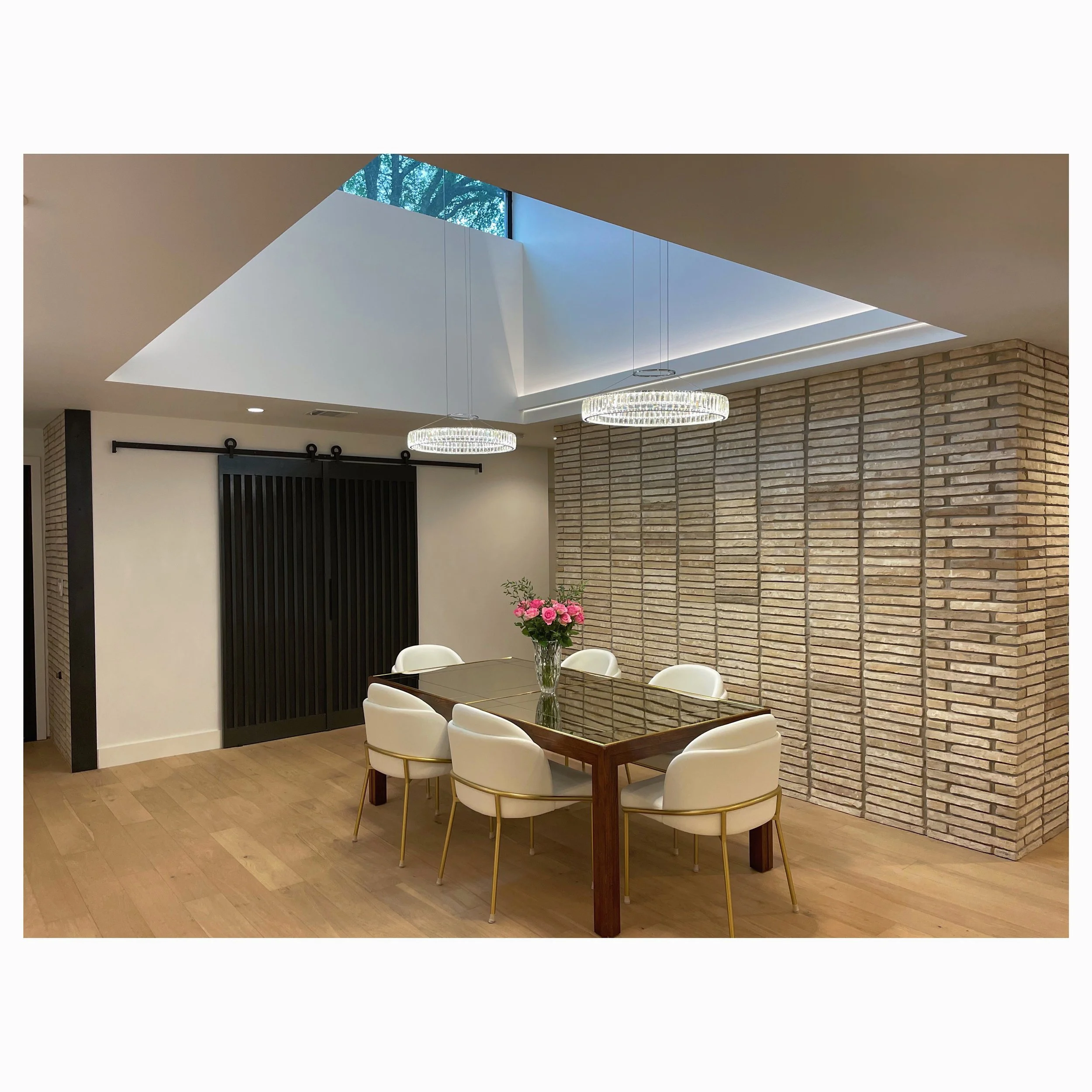 Modern dining room with a glass-topped wooden table, six white chairs, a vase with pink roses, brick accent wall, black sliding barn door, and skylight with contemporary chandeliers overhead.