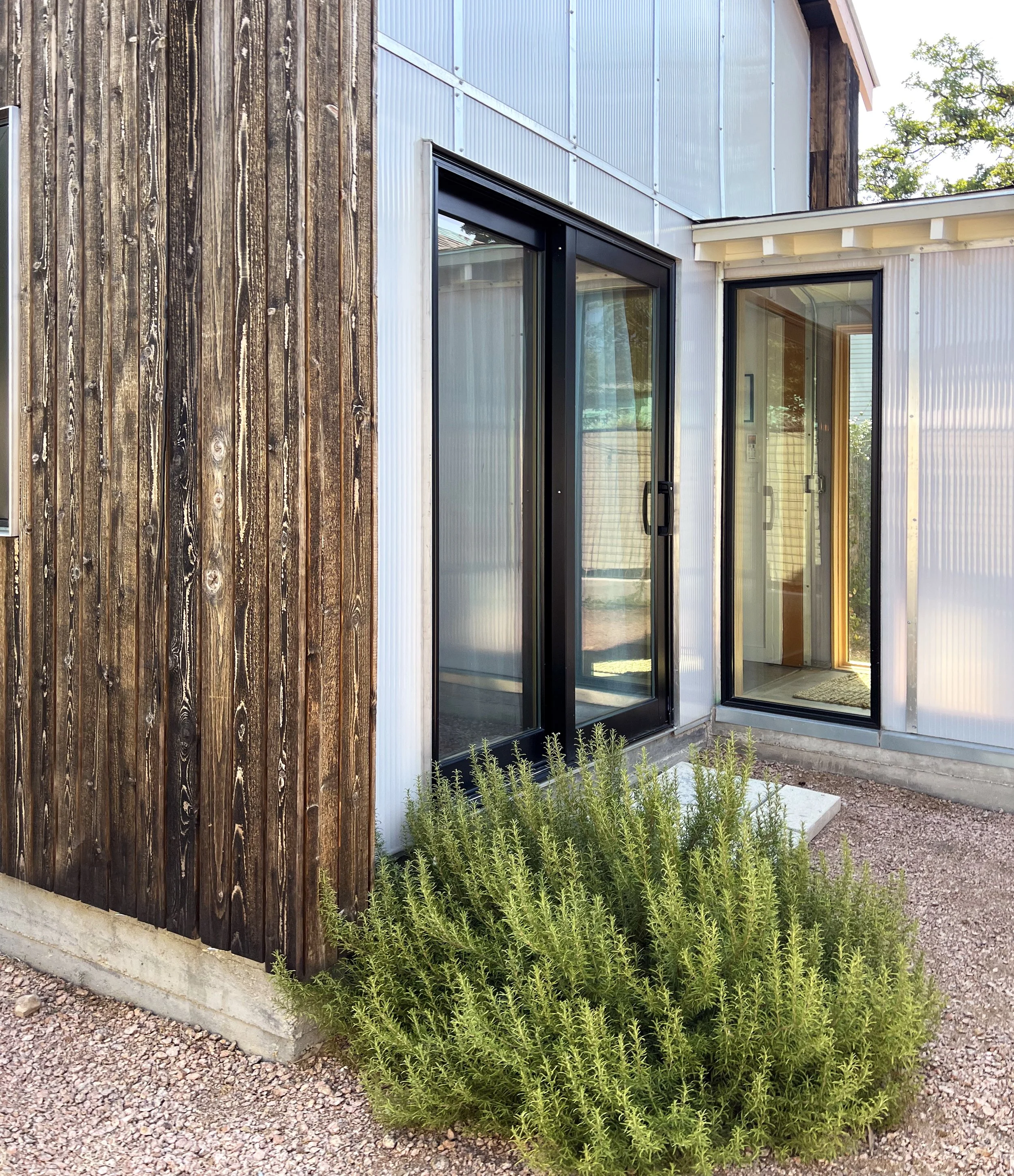 Exterior view of a modern building with a combination of wood and metal siding, featuring two glass sliding doors and a green shrub in the foreground, on a gravel ground.