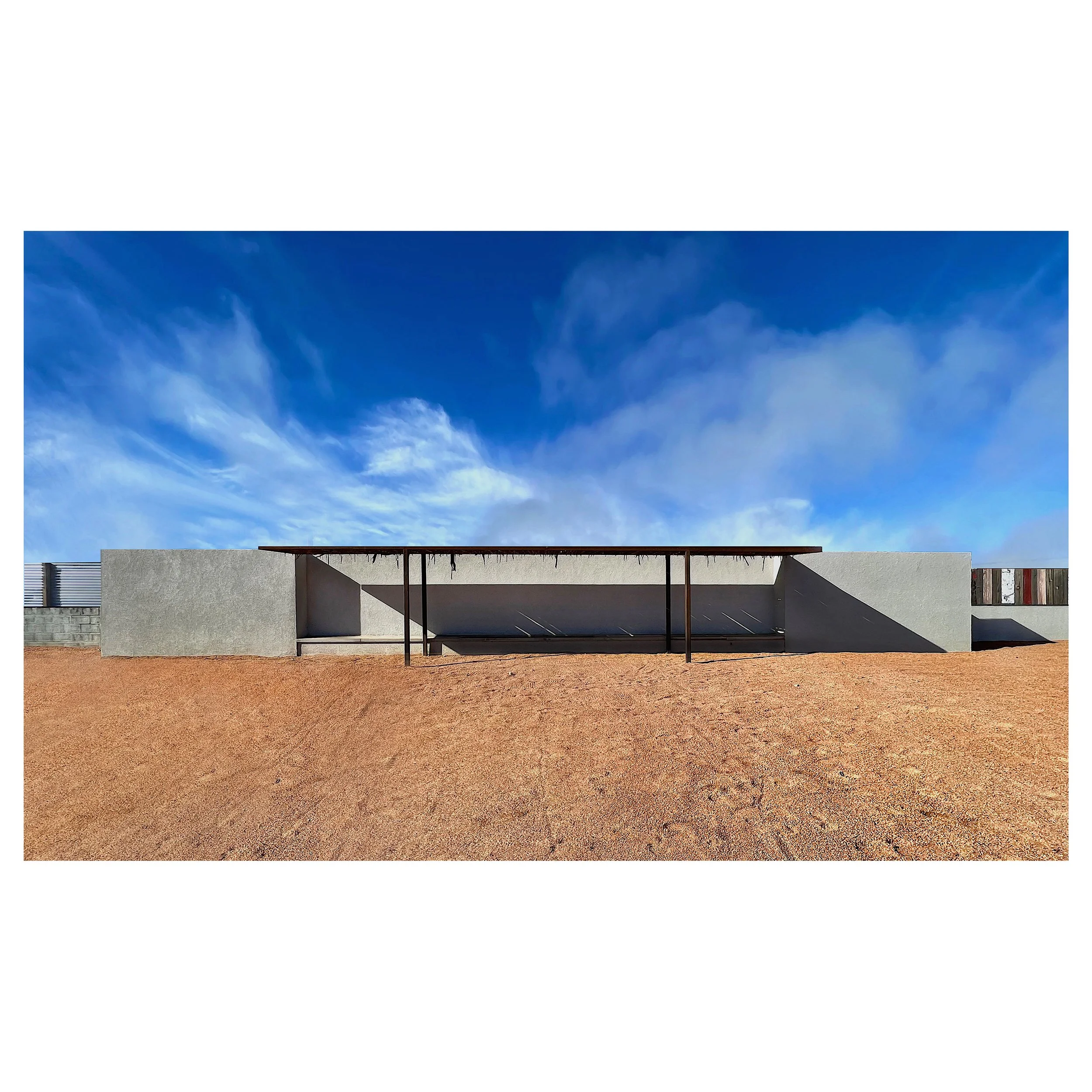 Modern concrete building with a cantilevered roof, set against a blue sky with wispy clouds, and a reddish dirt ground in the foreground.