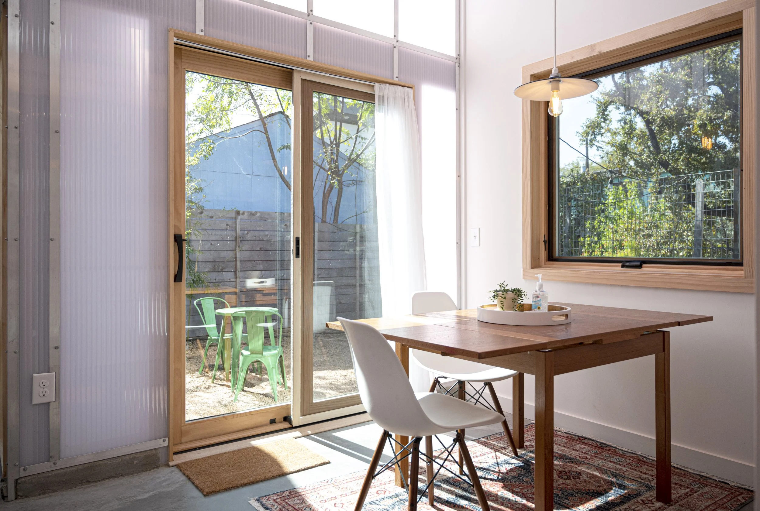 A bright dining area with a wooden table and two white chairs, a sliding glass door leading to a small outdoor patio with green plastic chairs, and a large window showing trees outside.