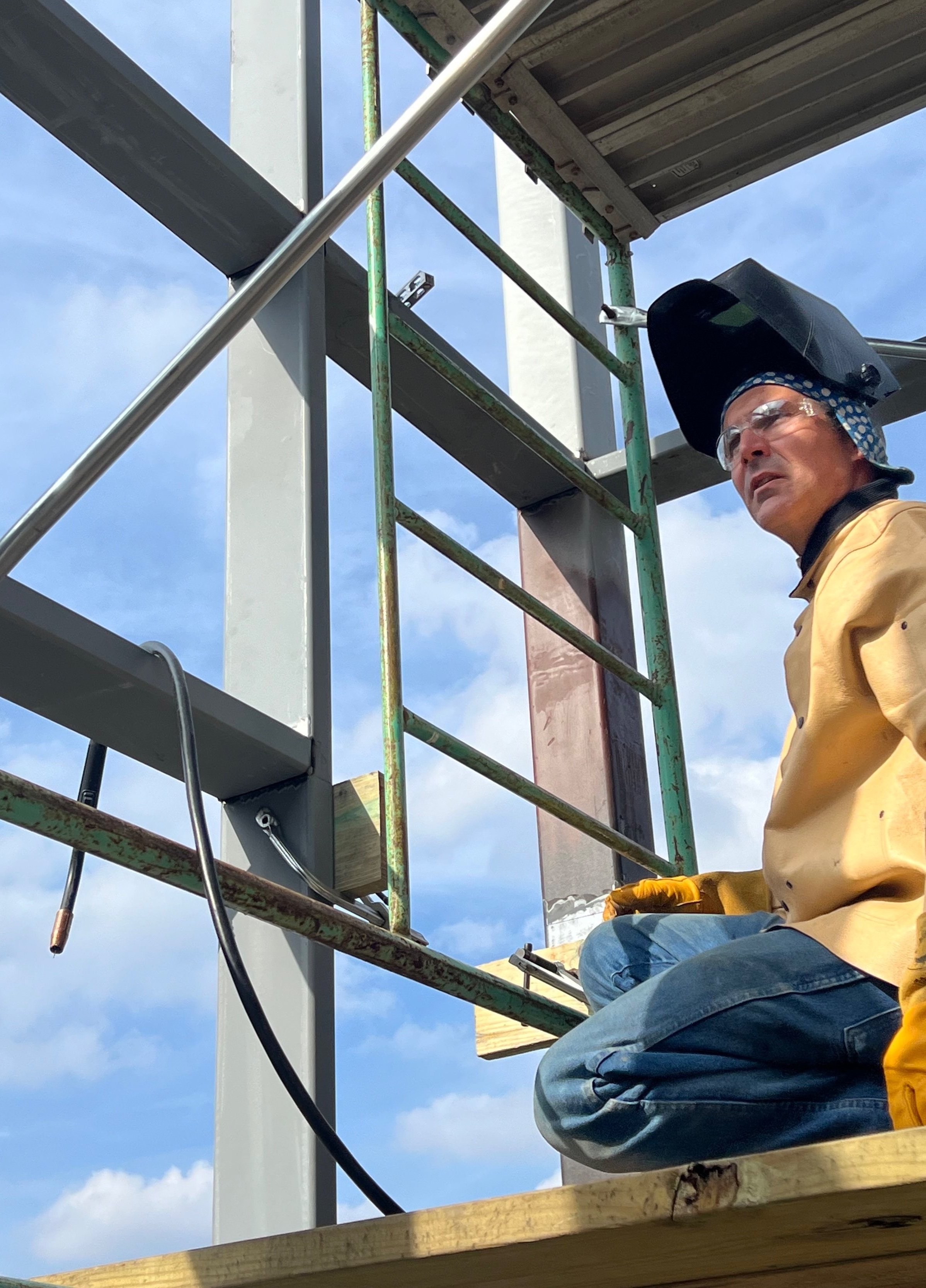 A construction worker wearing a welding helmet, safety glasses, yellow work jacket, and gloves, kneeling on a wooden platform at a construction site with steel and scaffolding around him, looking up at the structure.