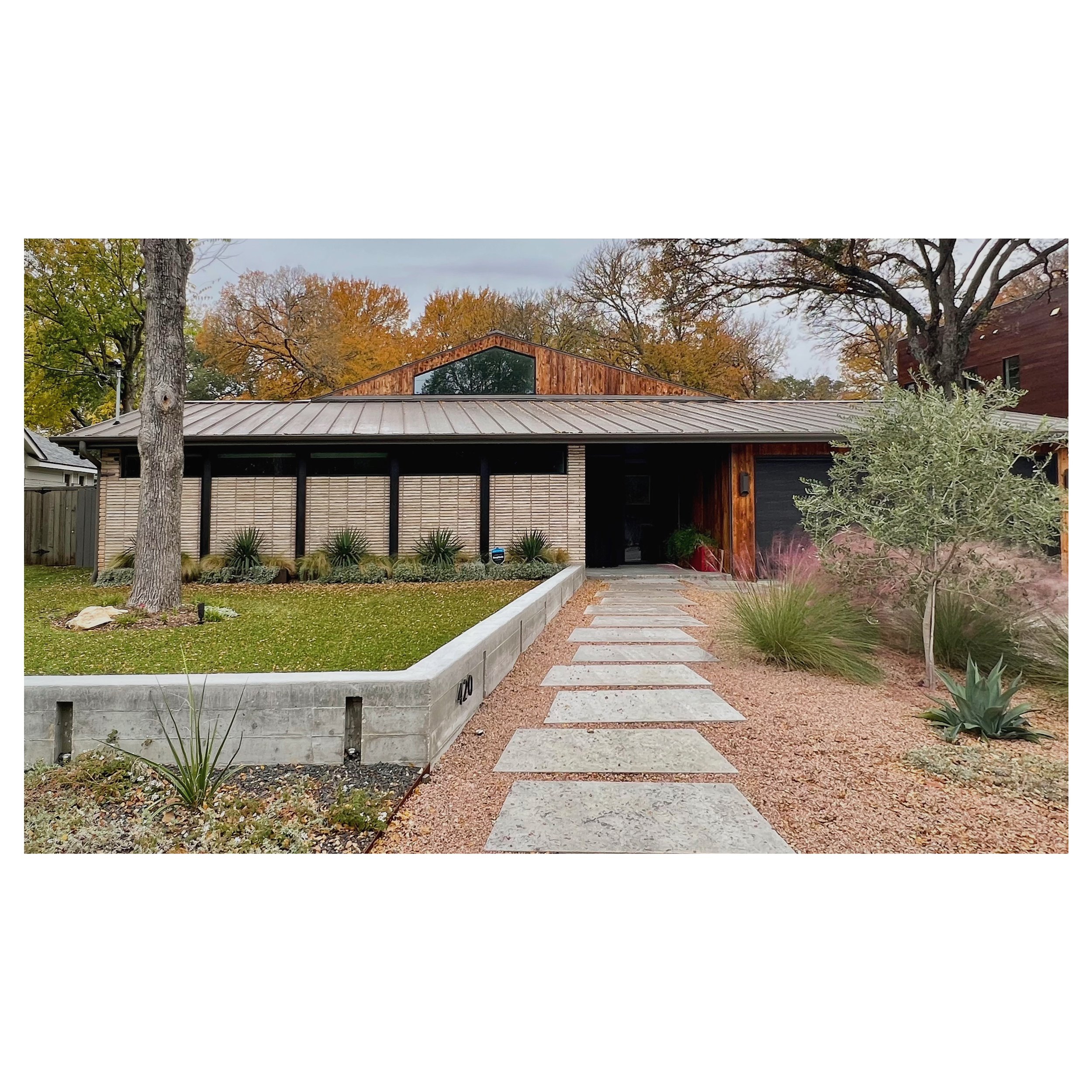 Modern house with wooden and brick exterior, metal roof, pathway with stepping stones, desert landscaping with succulents and trees.