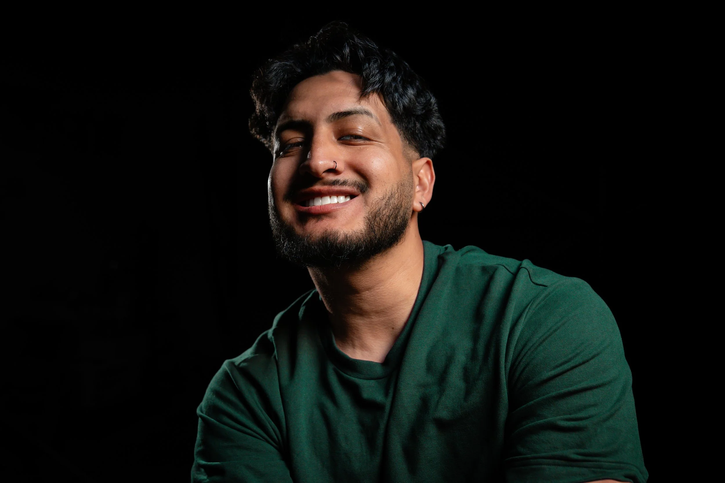 Smiling man with dark hair, beard, earrings, and nose stud, wearing a green shirt against a black background.
