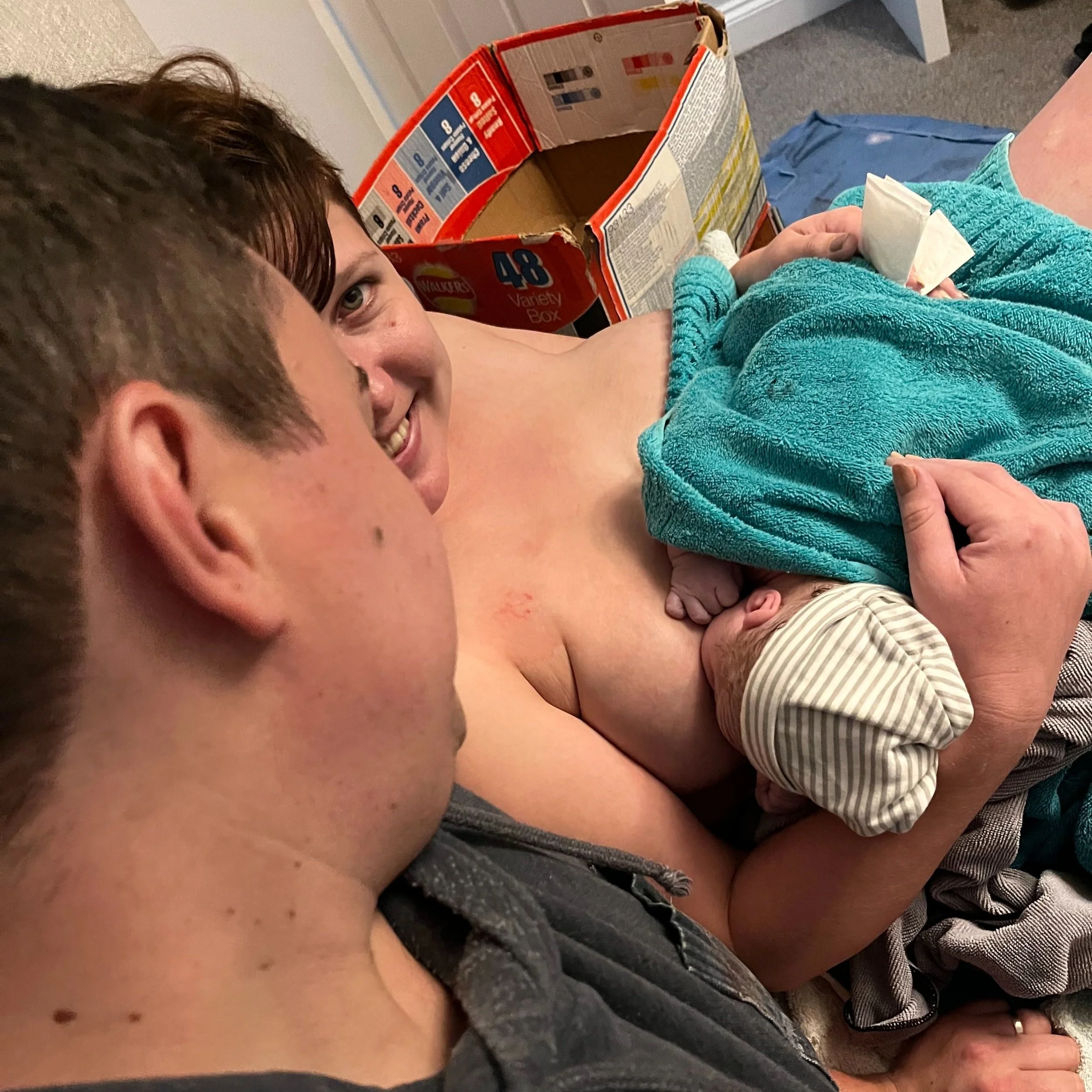 a newborn has his first feed while wrapped in a towel. mum and dad look on proudly
