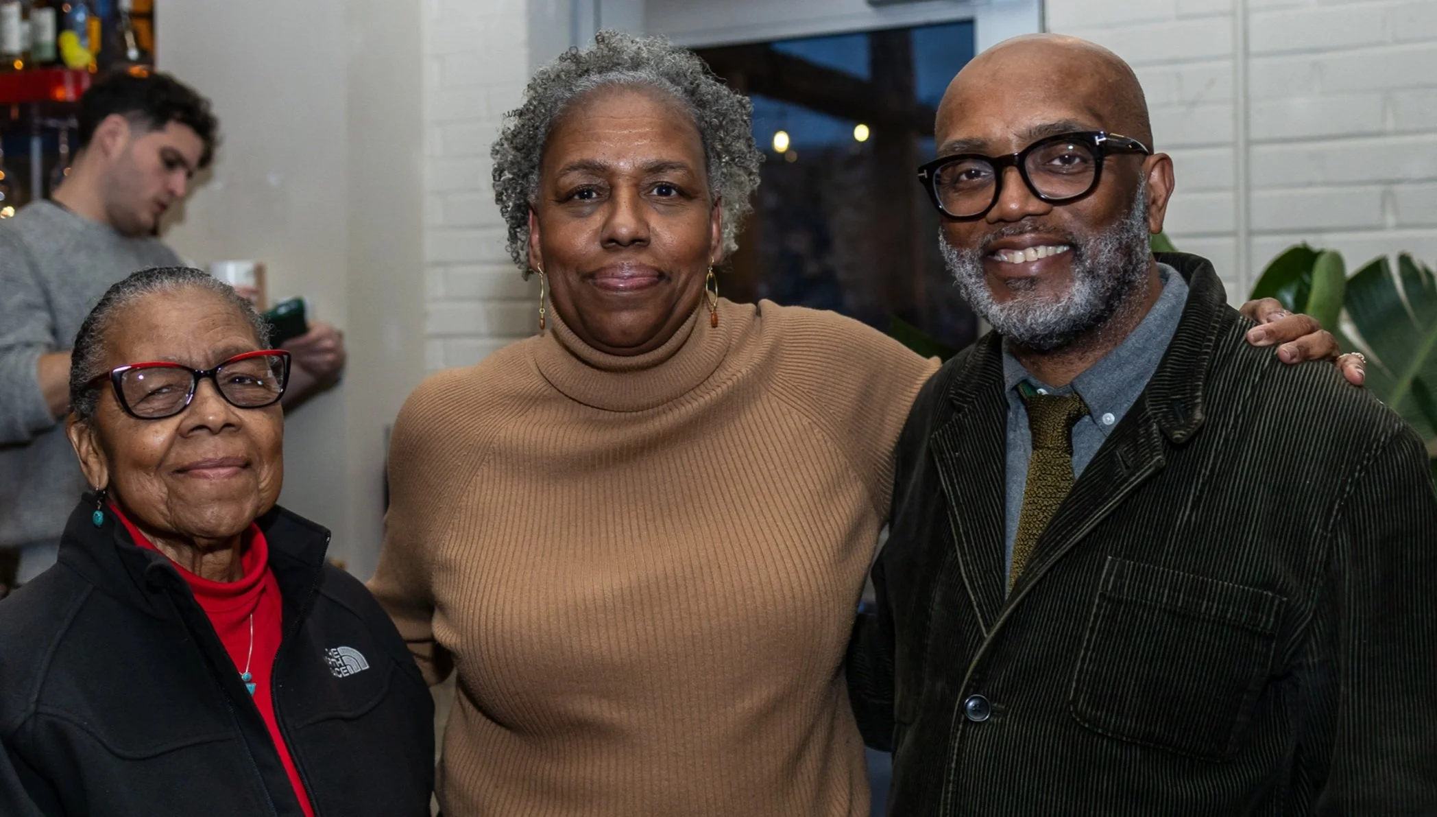 three Black family members stand together with their arms around eachother. On one end is the family matriarch, in the middle is her grown daughter, and on the other end is her daughter's husband