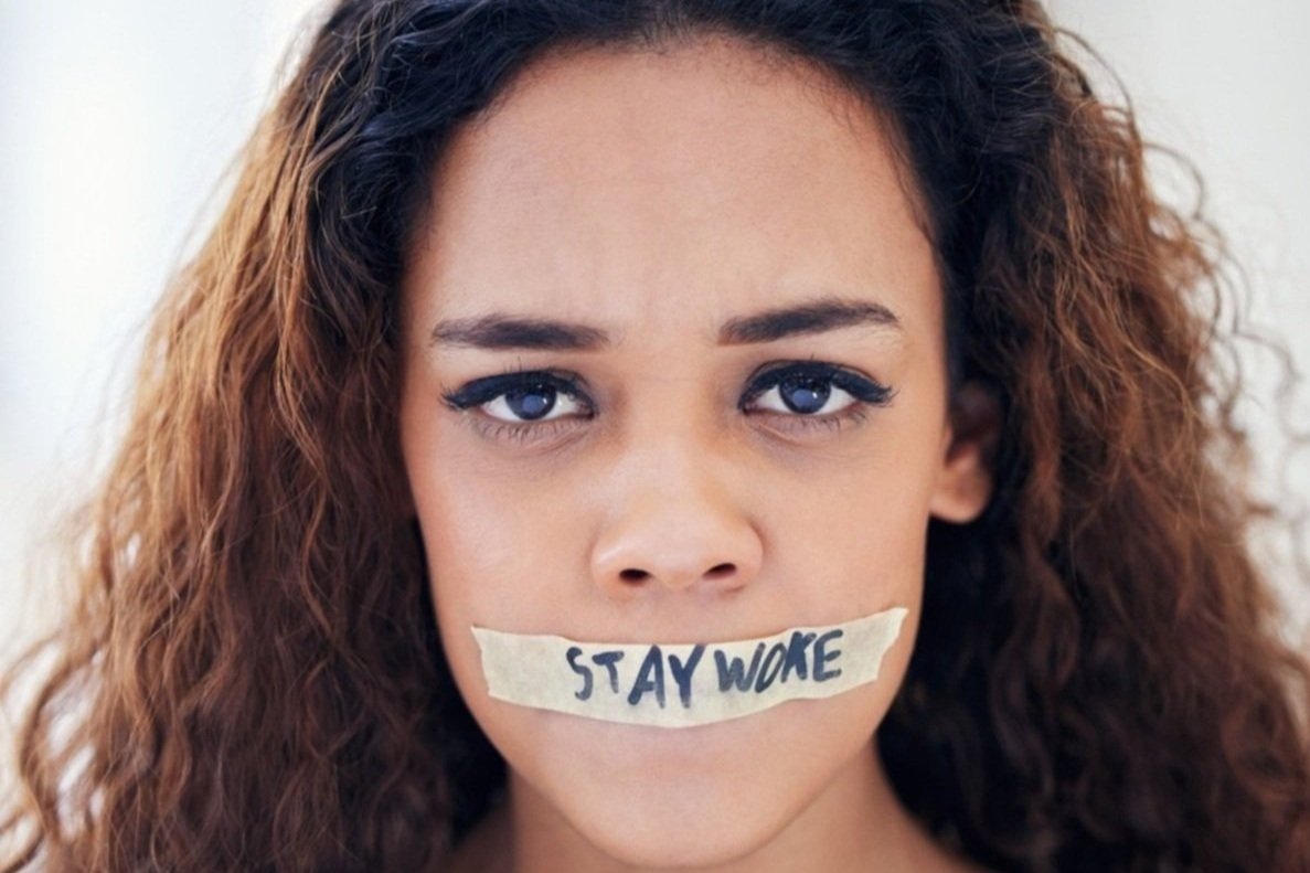 woman with long curly hair looks at the camera with masking tape over her mouth with the words "STAY WOKE" written on the tape in black marker