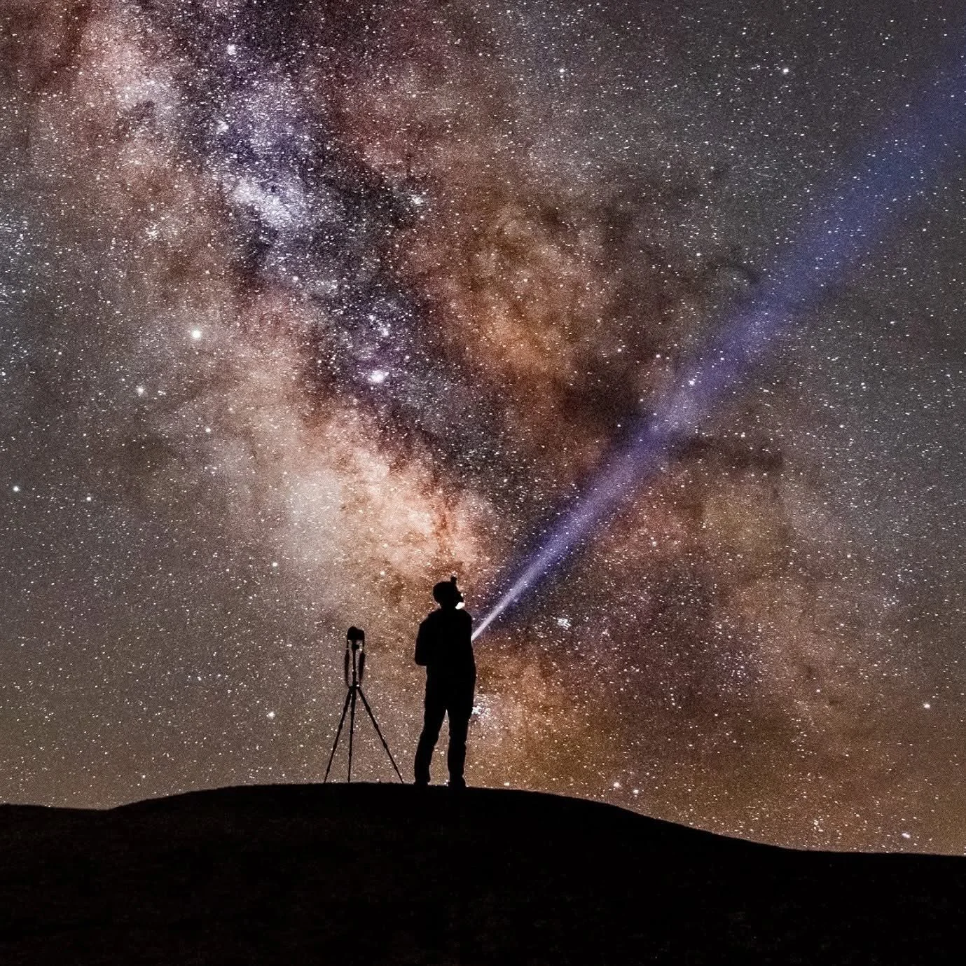 En person som står på en bergstopp med en ficklampa riktad mot stjärnorna och Vintergatan över himlen, med ett trebenigt stativ bredvid.